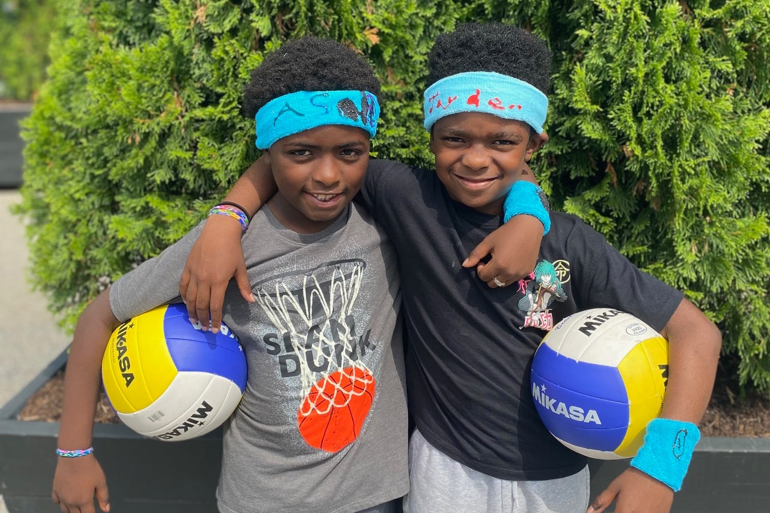 Two boys in blue sweatbands stand next to each other holding volleyballs.