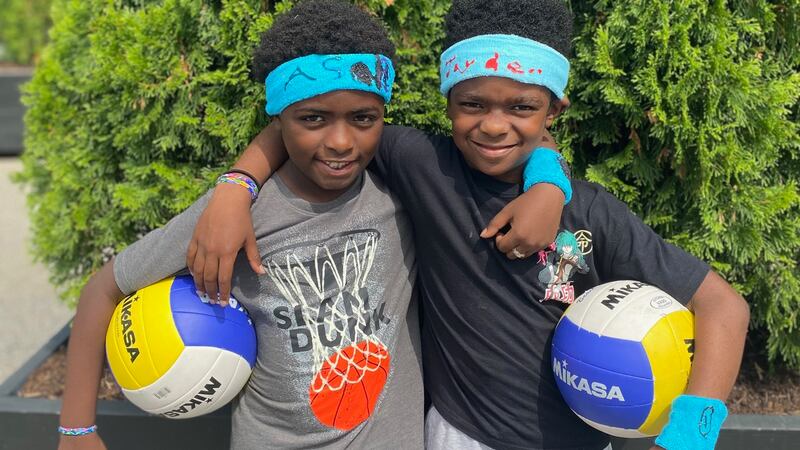 Two boys in blue sweatbands stand next to each other holding volleyballs.