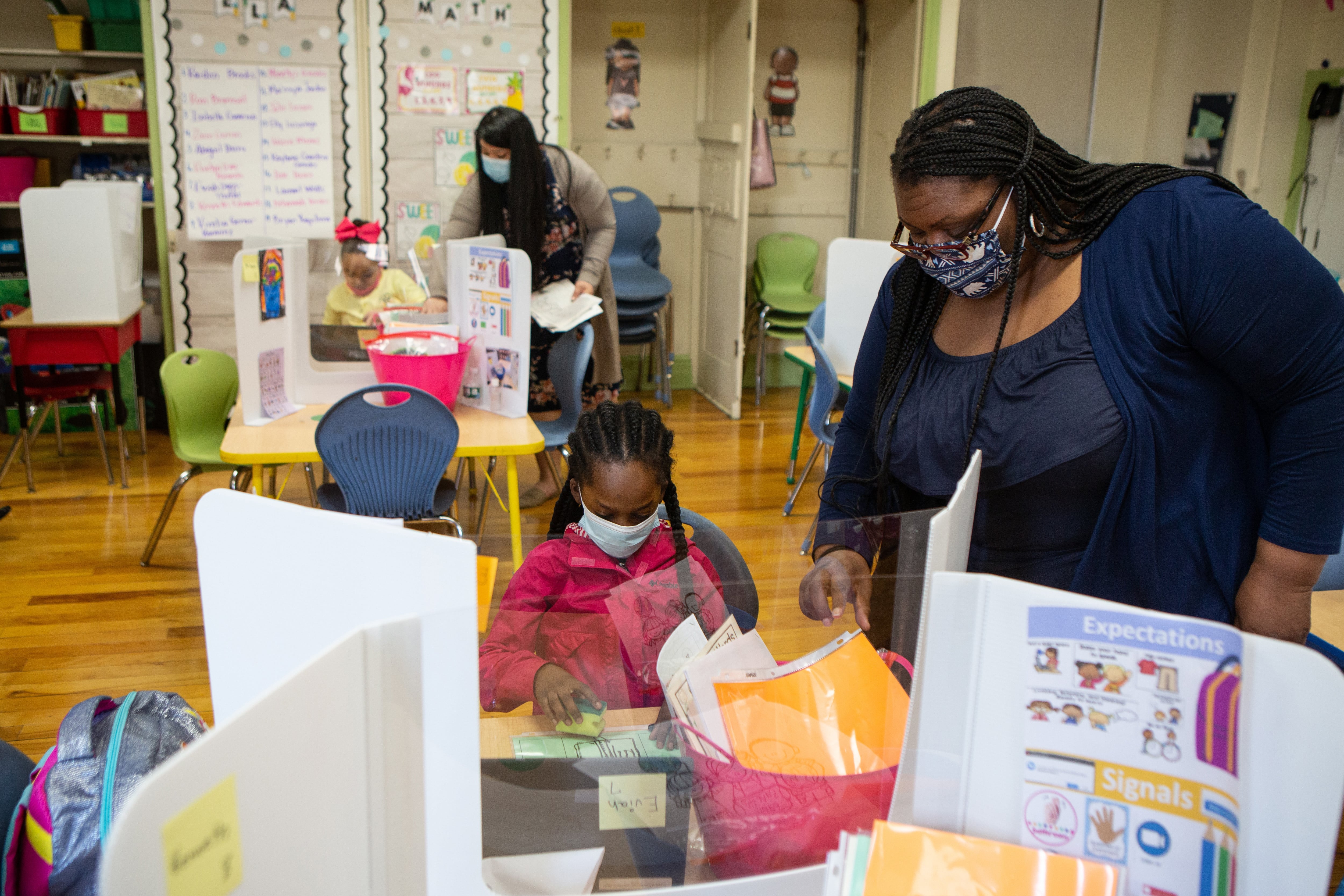 A teacher works with a student at a table in a classroom. Both are wearing masks.