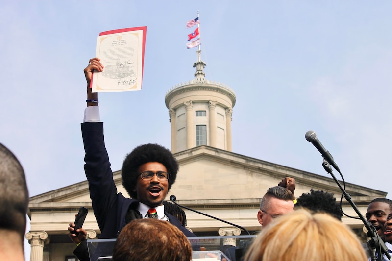 A man with glasses and an Afro style haircut stands with his hand in the air holding a piece of paper.