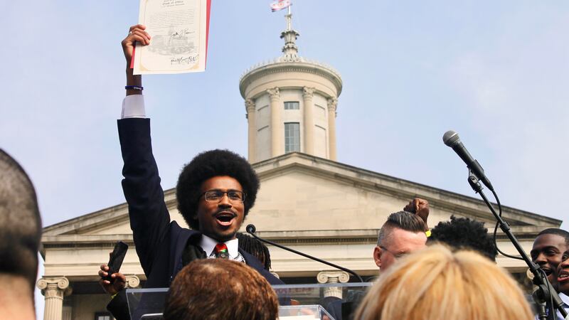 A man with glasses and an Afro style haircut stands with his hand in the air holding a piece of paper.