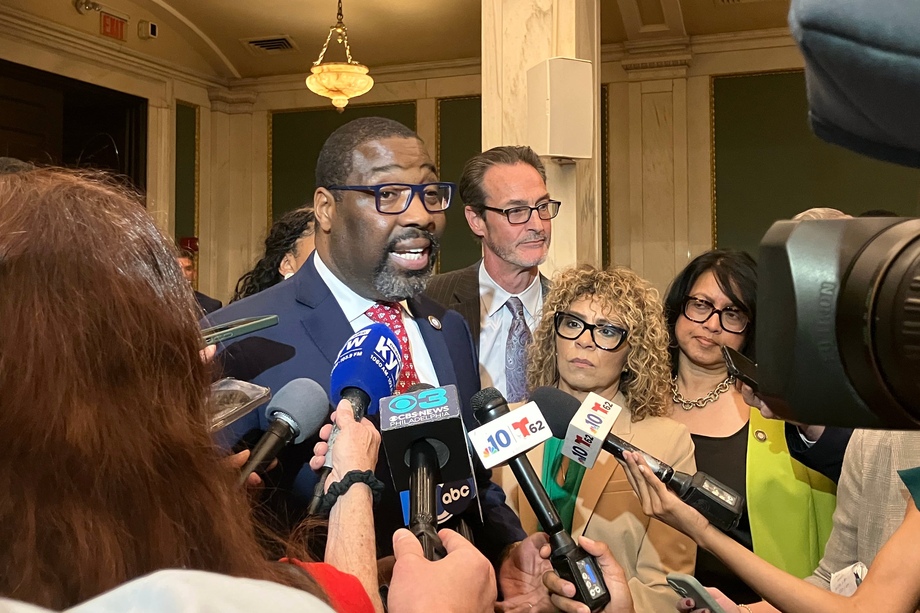 A photograph of a Black man wearing glasses and a suit surrounded by members of the press holding microphones in the lobby of a building.