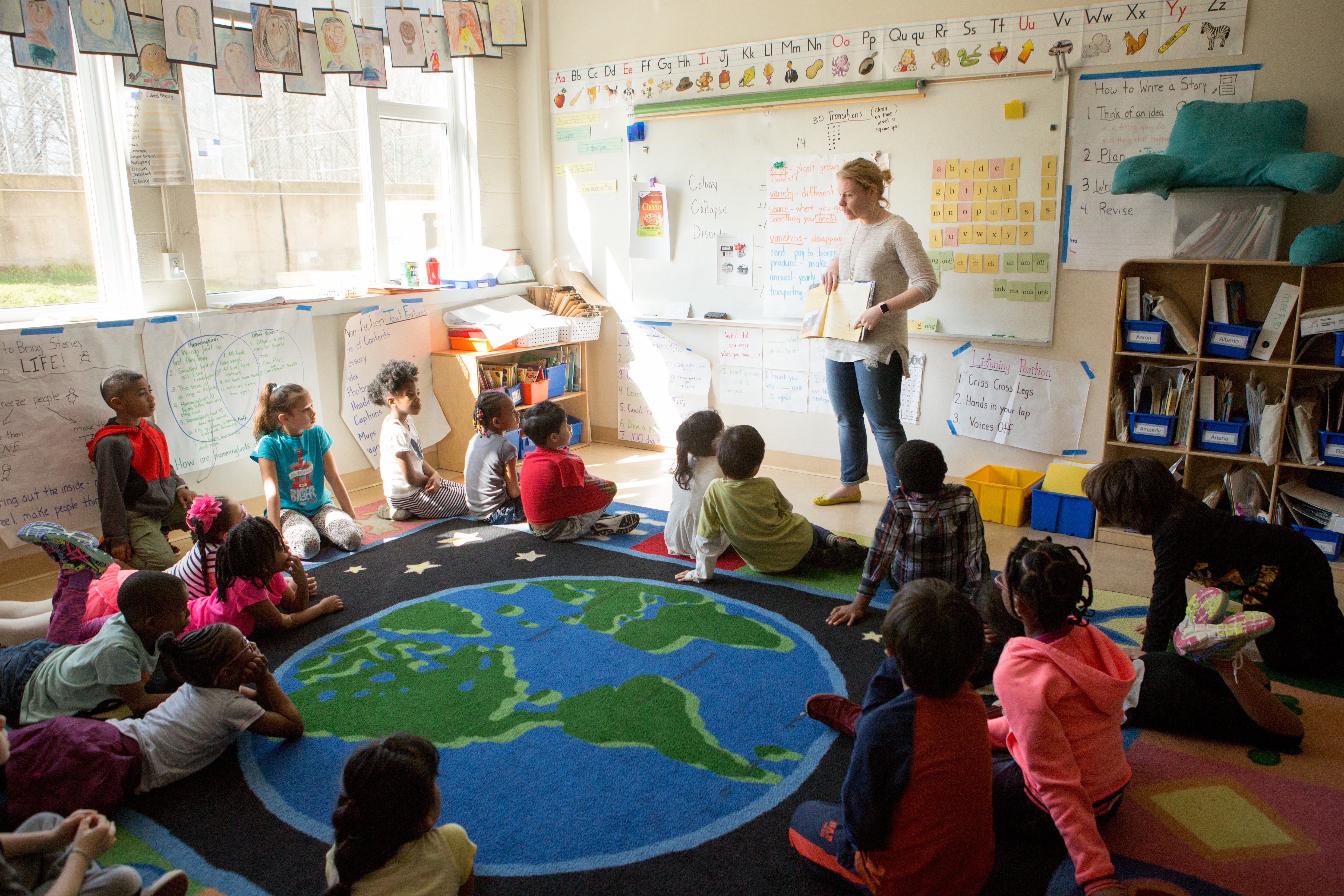 A group of children sit on a rug with a drawing of the Earth on it as their teacher, wearing a white sweater and blue jeans, conducts a lesson in front of a white board.