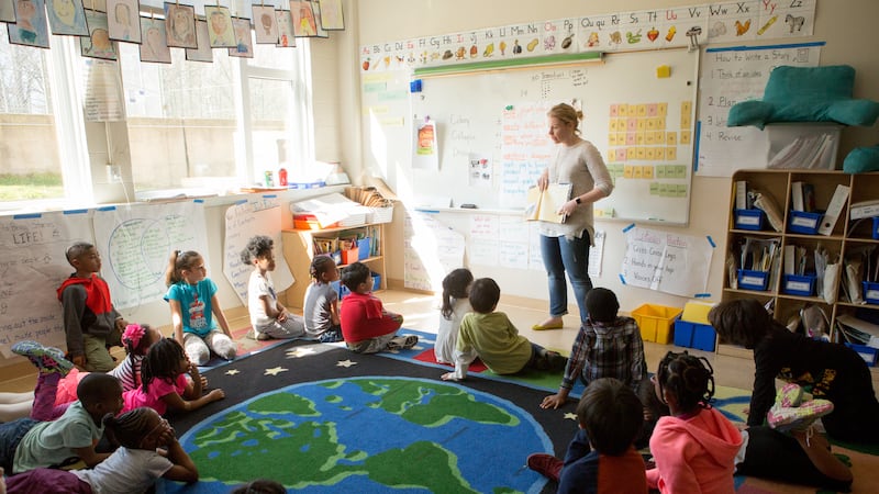A group of children sit on a rug with a drawing of the Earth on it as their teacher, wearing a white sweater and blue jeans, conducts a lesson in front of a white board.