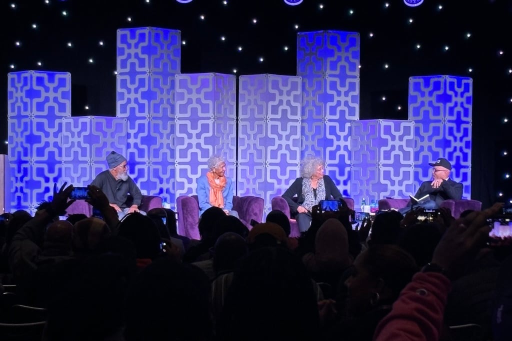 Four adults sit on a stage in front of a large crowd.