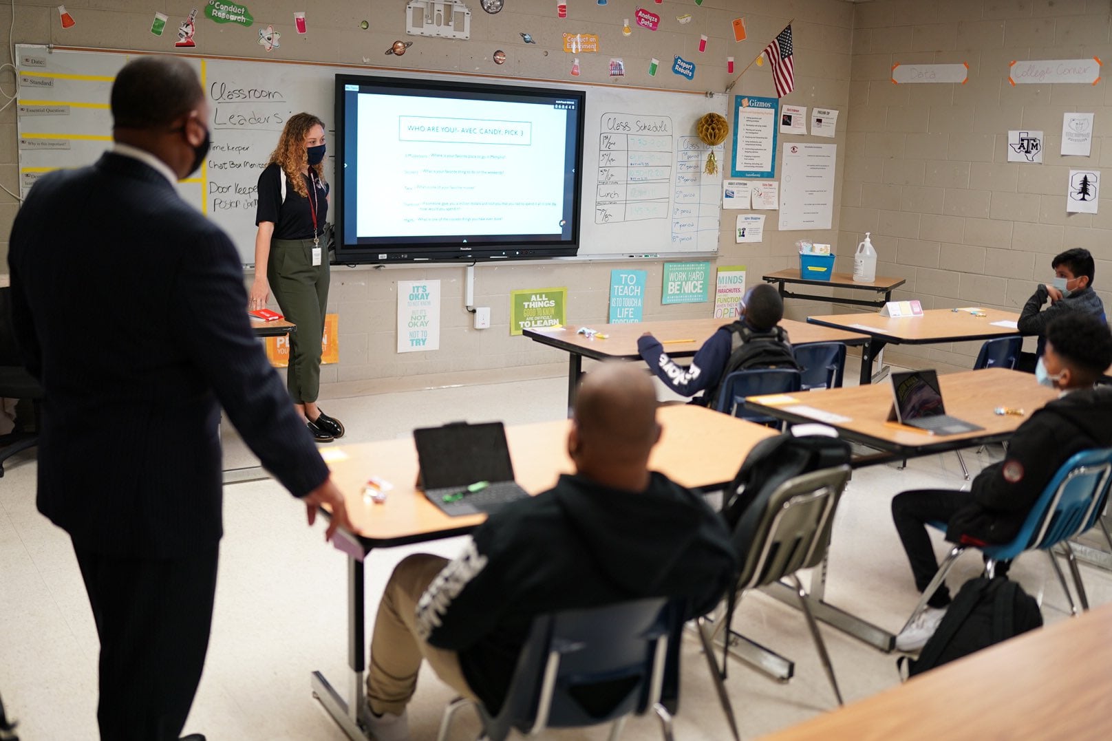 Memphis Superintendent Joris Ray stands next to a student’s desk while a teacher instructs from the front of the room with a mask on