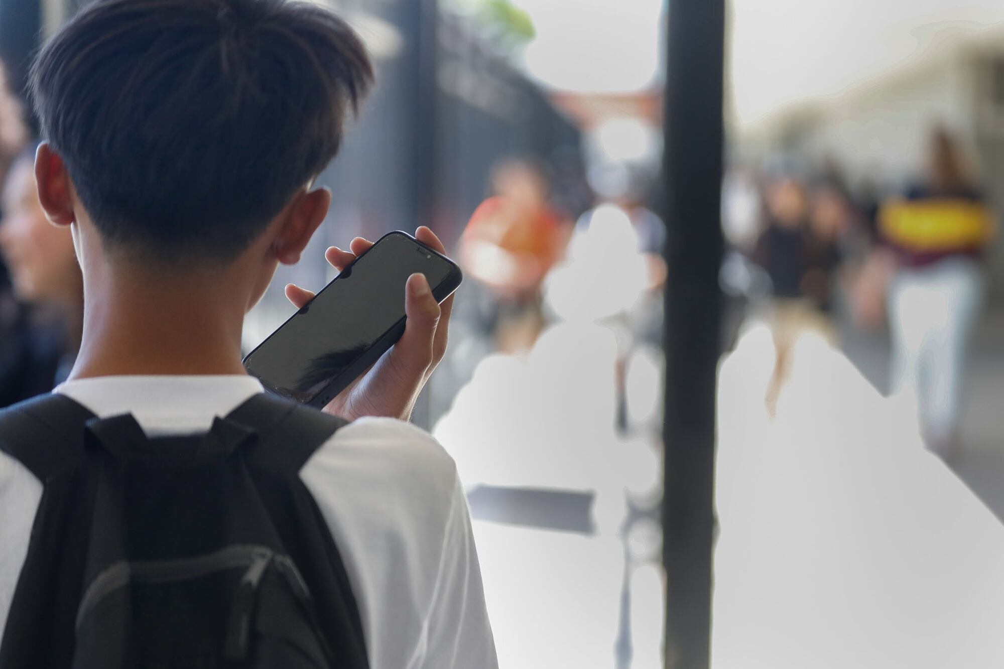 A view from behind a middle or high school student with short hair and wearing a backpack. The student is holding up a cellphone while walking outside of a school building.