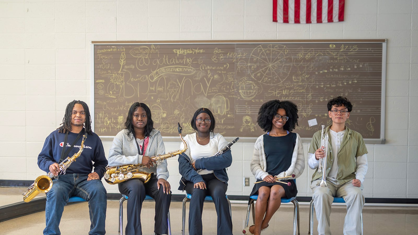 Five students, most holding musical instruments, sit in front of a blackboard.
