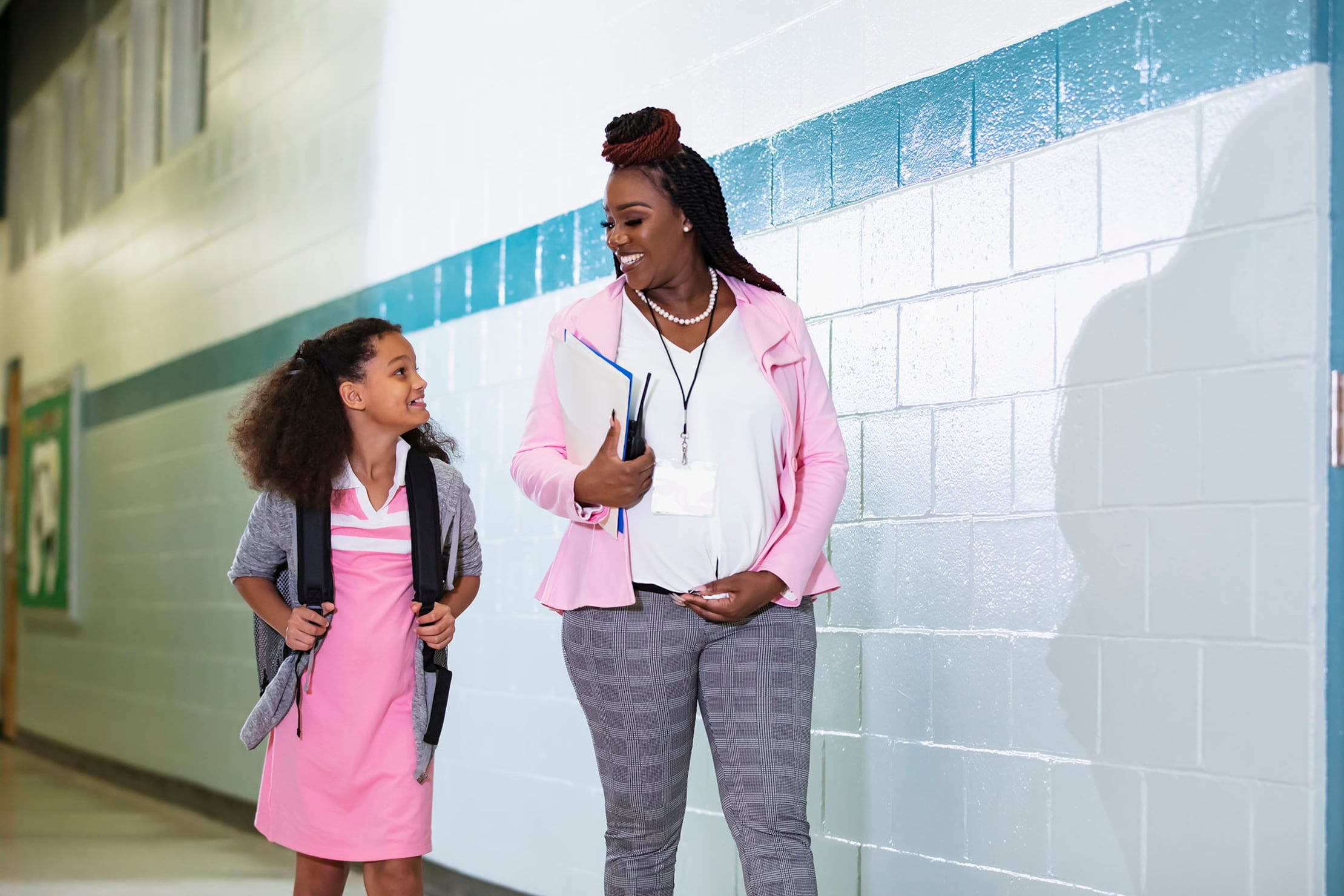 A photograph of a Black pregnant teacher walking alongside a young Black student in school hallway.