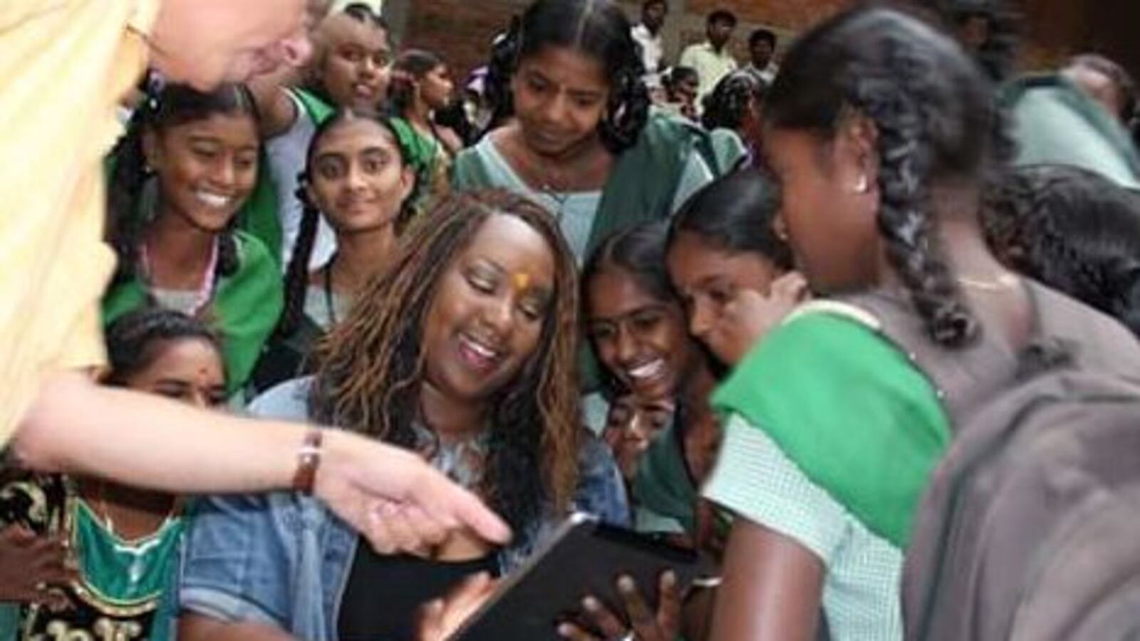 A teacher holds a tablet computer. A group of Indian children are gathered around her smiling. Melissa Collins visits with students during her two-week trip to India last summer.