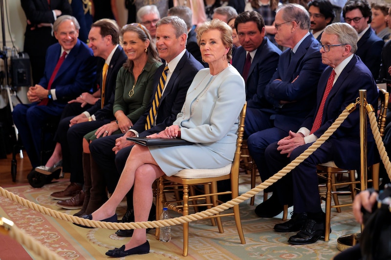 A white, older woman in a blue skirt suit sits on a chair of a corner of a few rows of chairs with various men and women in suits.