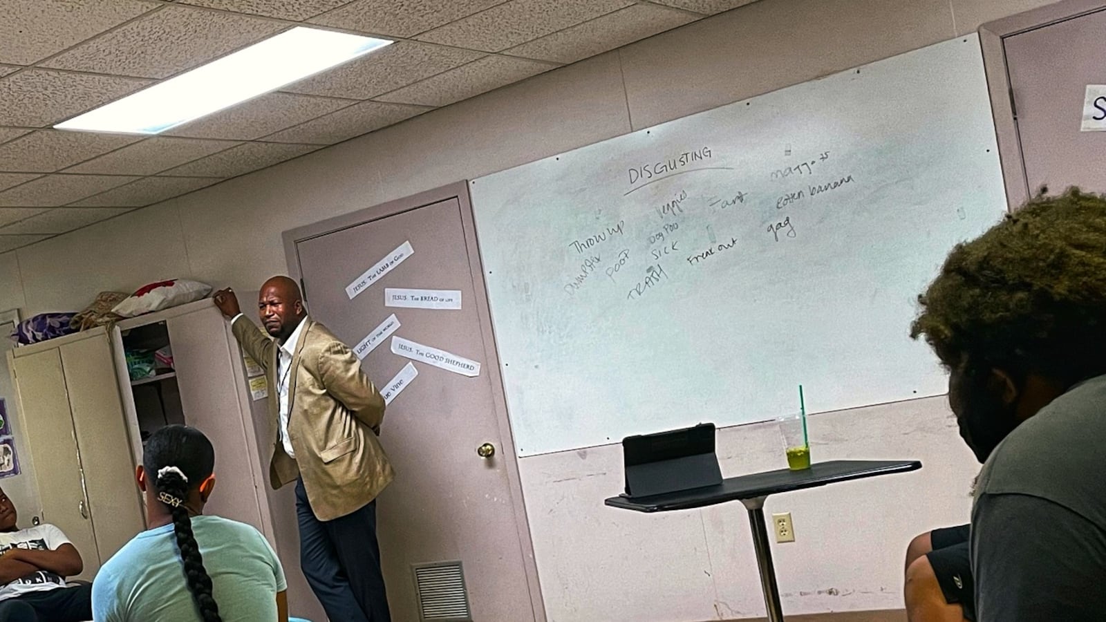 A well-dressed man in a khaki blazer and jeans stands near a screen in front of a class of teenage students.