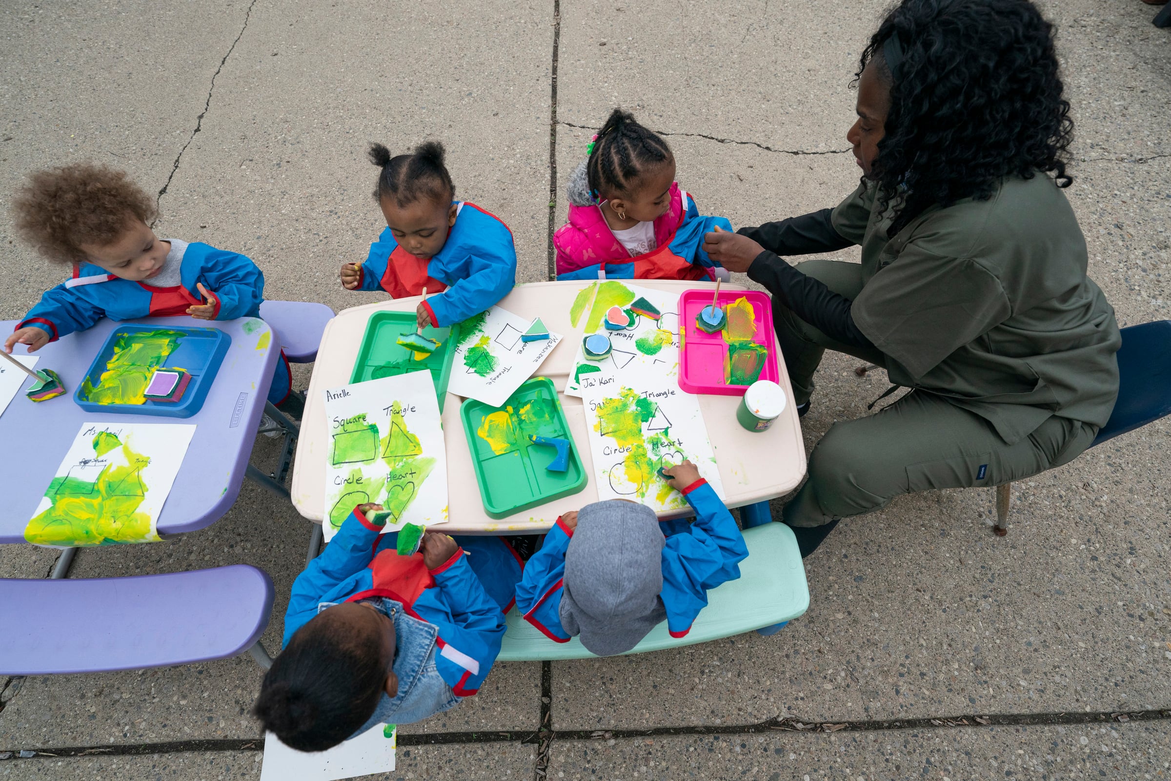 A photograph of an adult woman with long dark hair and wearing green scrubs sits at a small table next to five children.