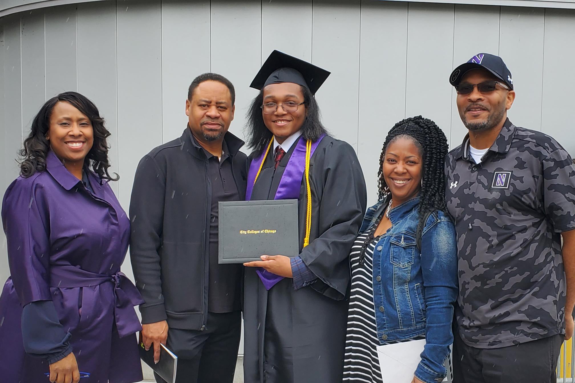 Two women and two men pose with a teen in a graduation cap and gown holding a diploma.