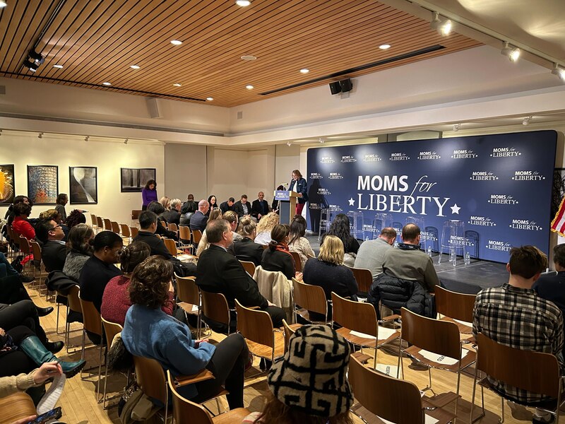 A woman stands at a podium with a "Moms for Liberty" sign behind her in front of a crowd of seated guests.