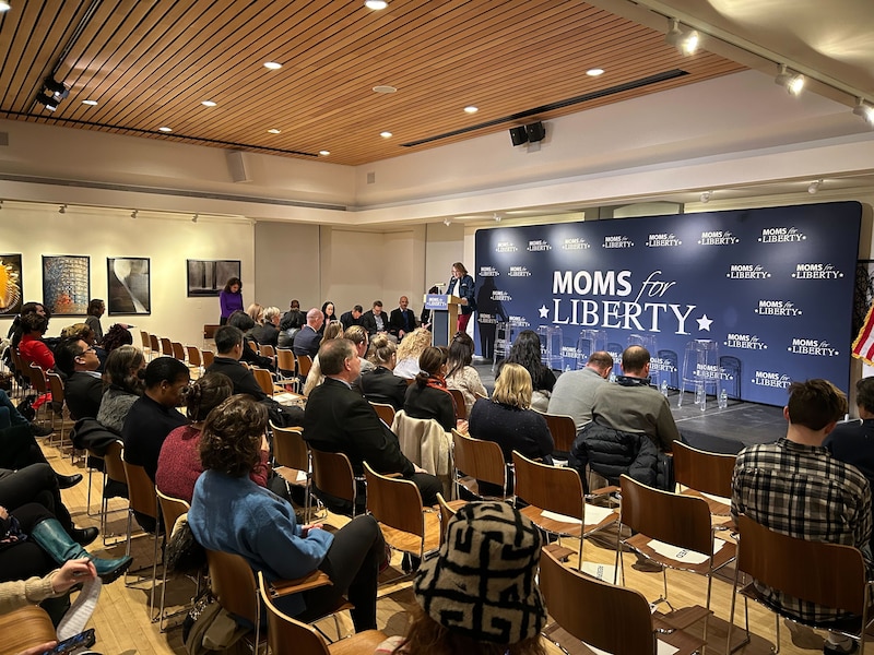 A woman stands at a podium with a "Moms for Liberty" sign behind her in front of a crowd of seated guests.