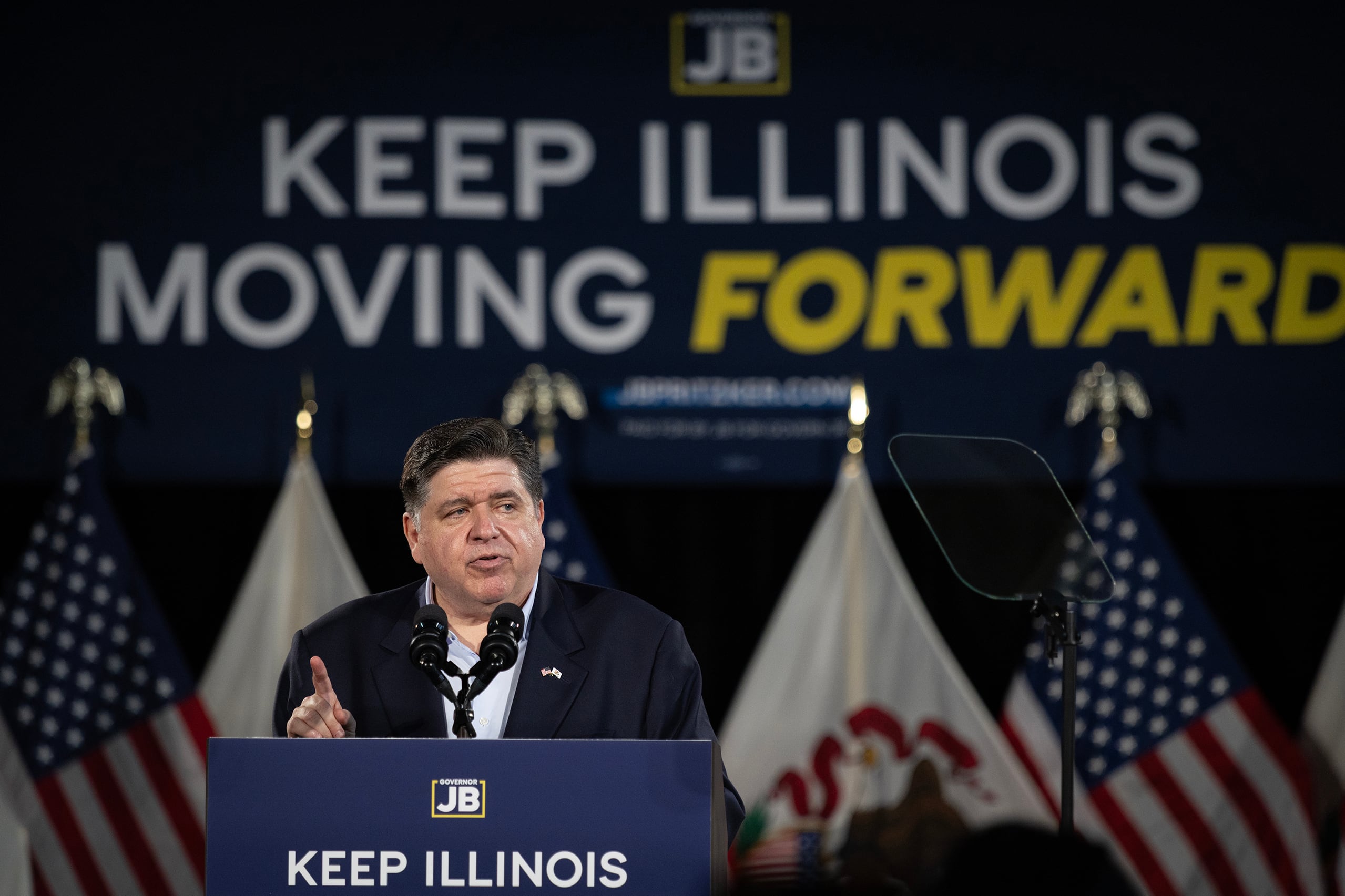 A photograph of a white man in a suit standing behind a podium and in front of a large backdrop with flags in the background.