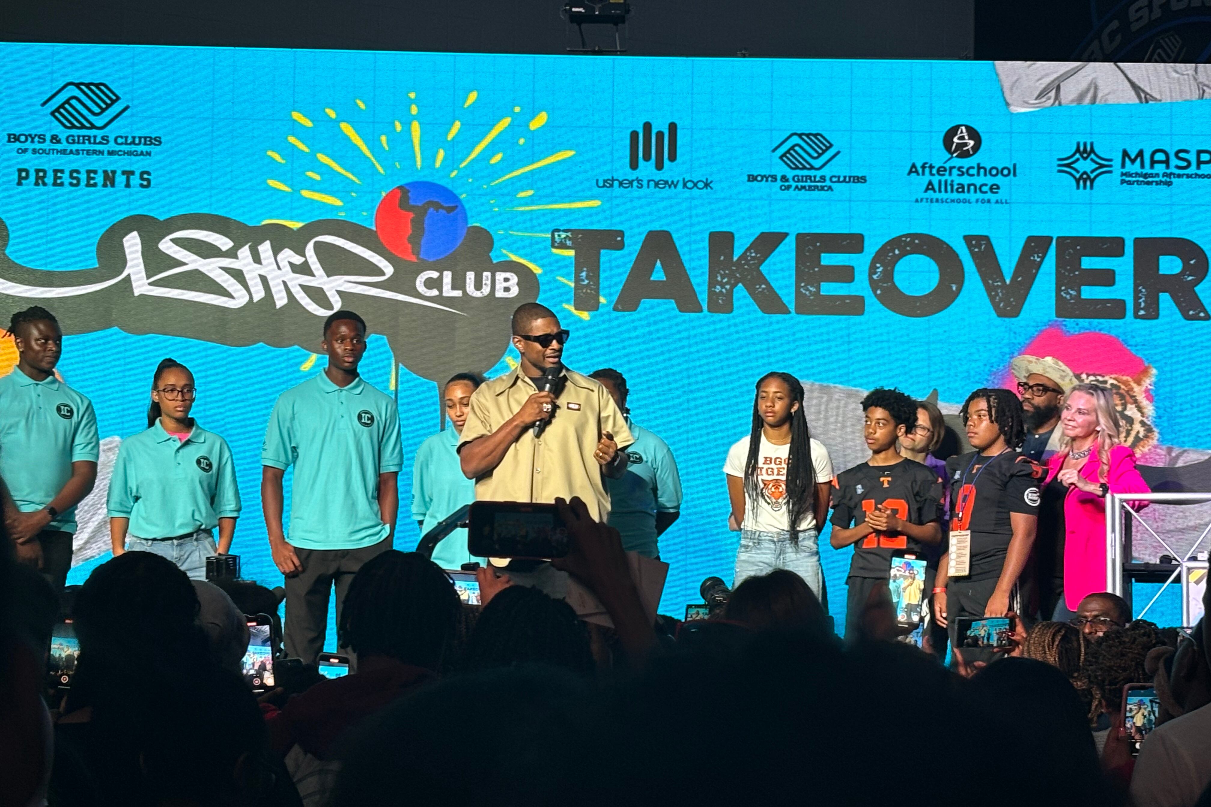 A man wearing sunglasses and a yellow shirt holds a microphone as he speaks to young people attending a Detroit event. Kids wearing colorful shirts stand behind him.
