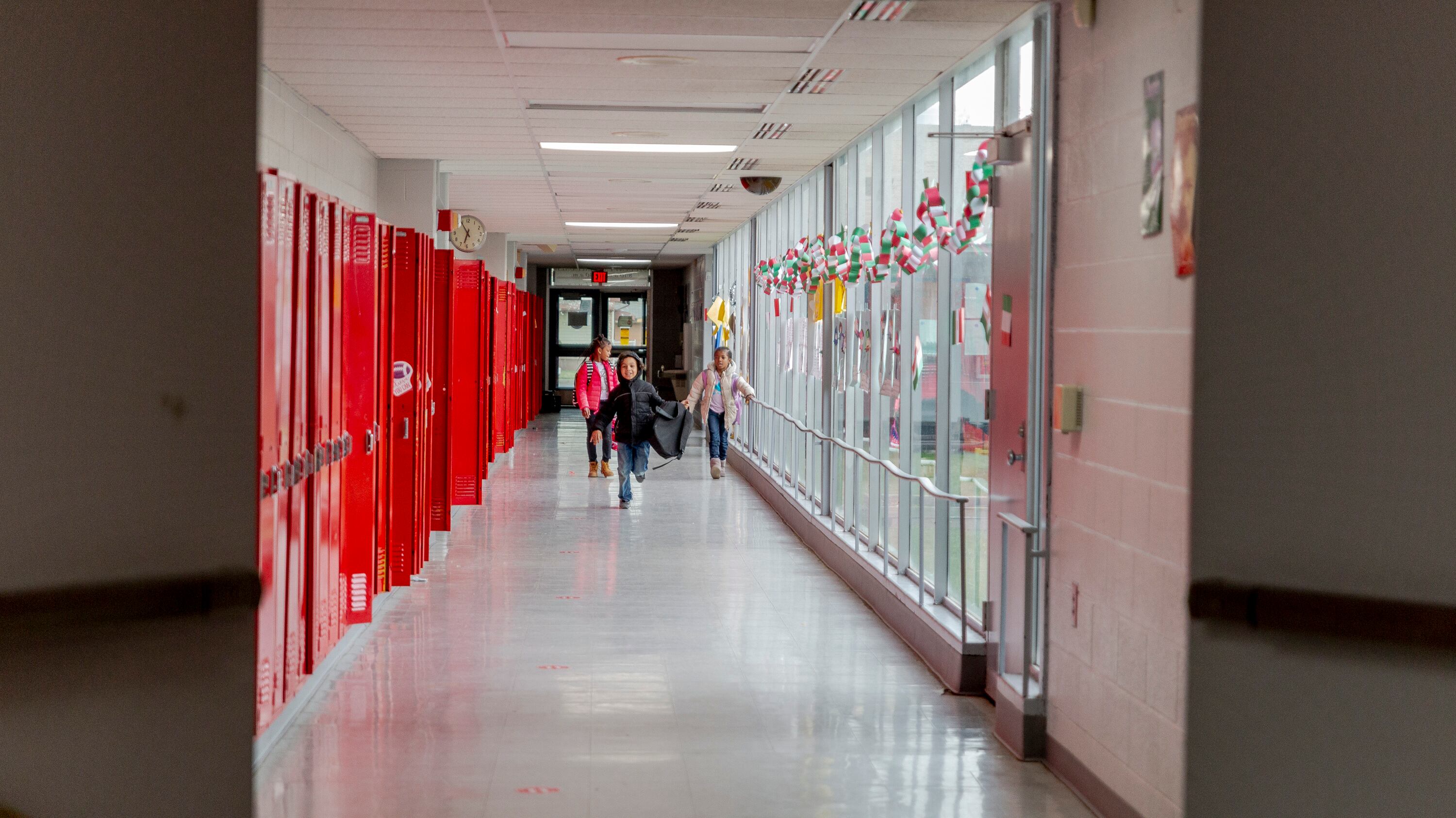 A view down a school hallway lined by red school lockers. The hallway is mostly empty, but a few students are visible in the distance.