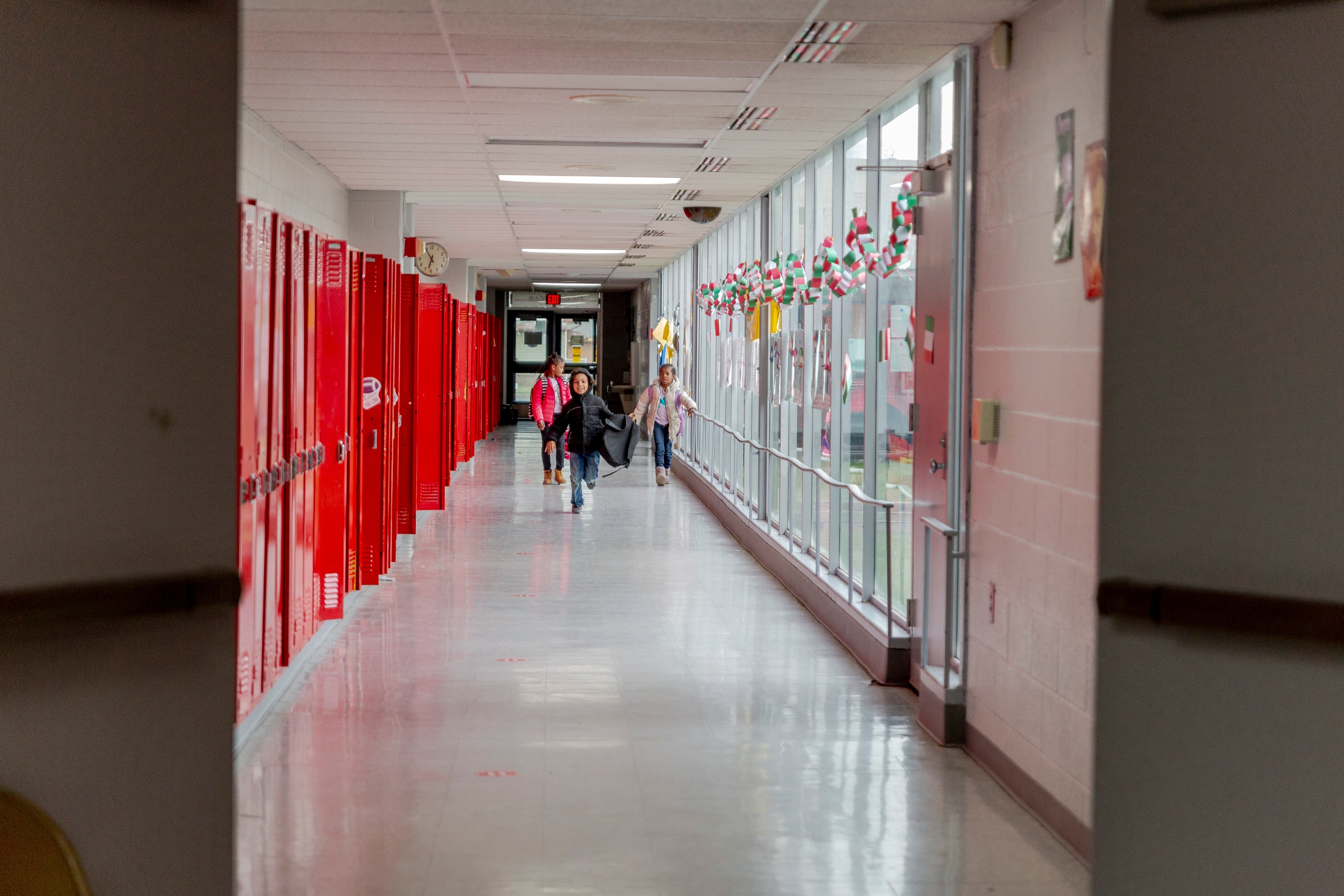 A view down a school hallway lined by red school lockers. The hallway is mostly empty, but a few students are visible in the distance.
