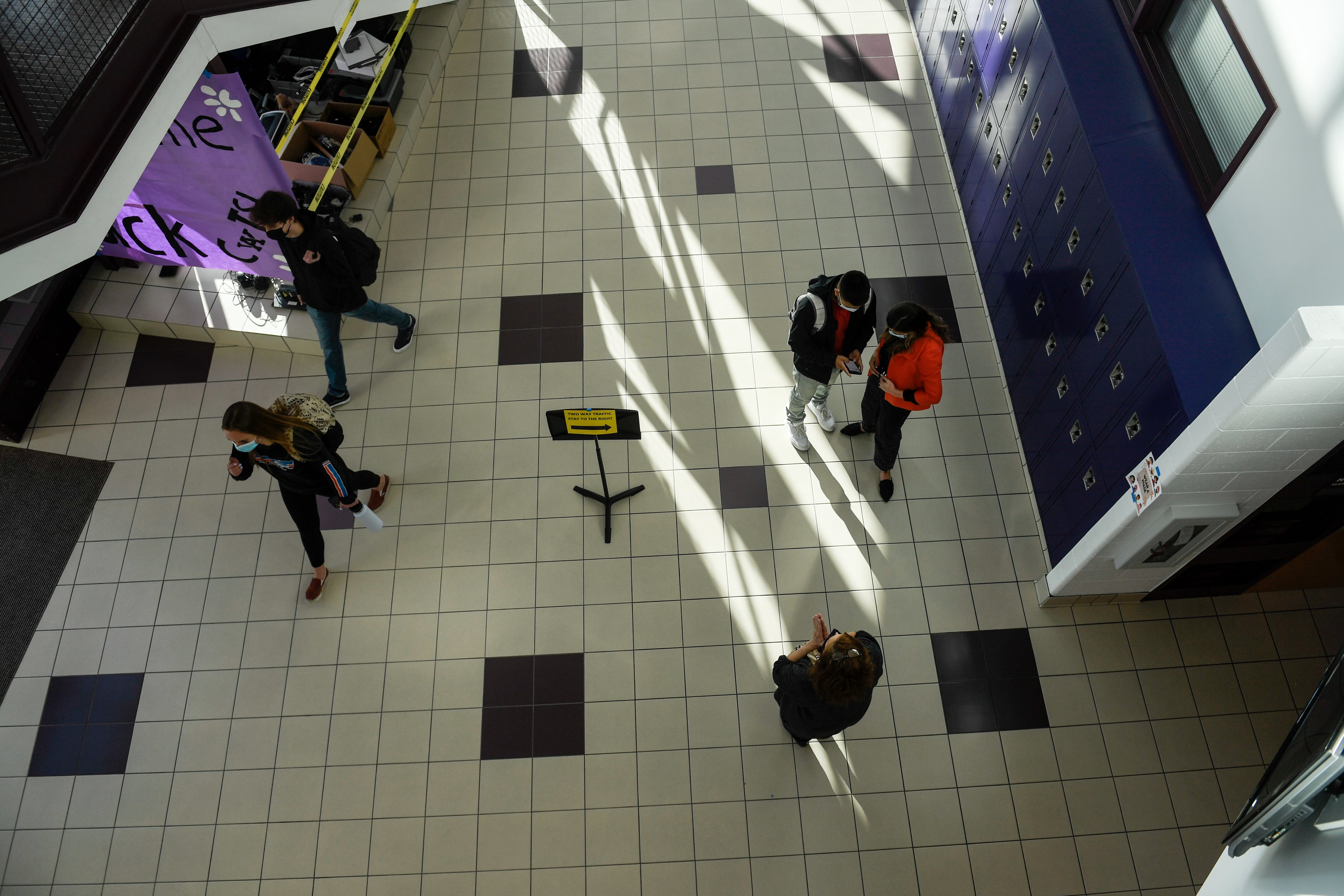 Seen from above, small groups of masked students walk in a wide school hallway.