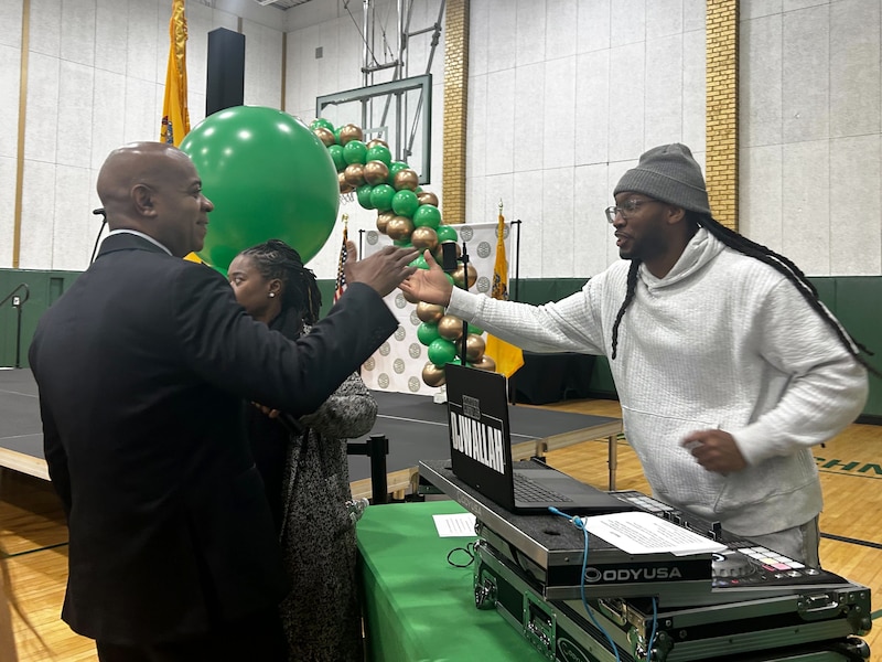 A Black man in a suit greets a Black man in a white sweater with a DJ set and a laptop in a school gym.
