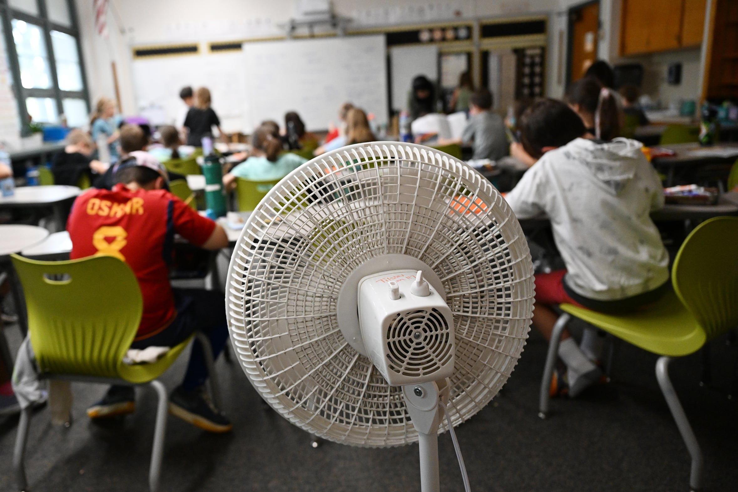 A white fan points toward the backs of students sitting at their desks in a classroom.