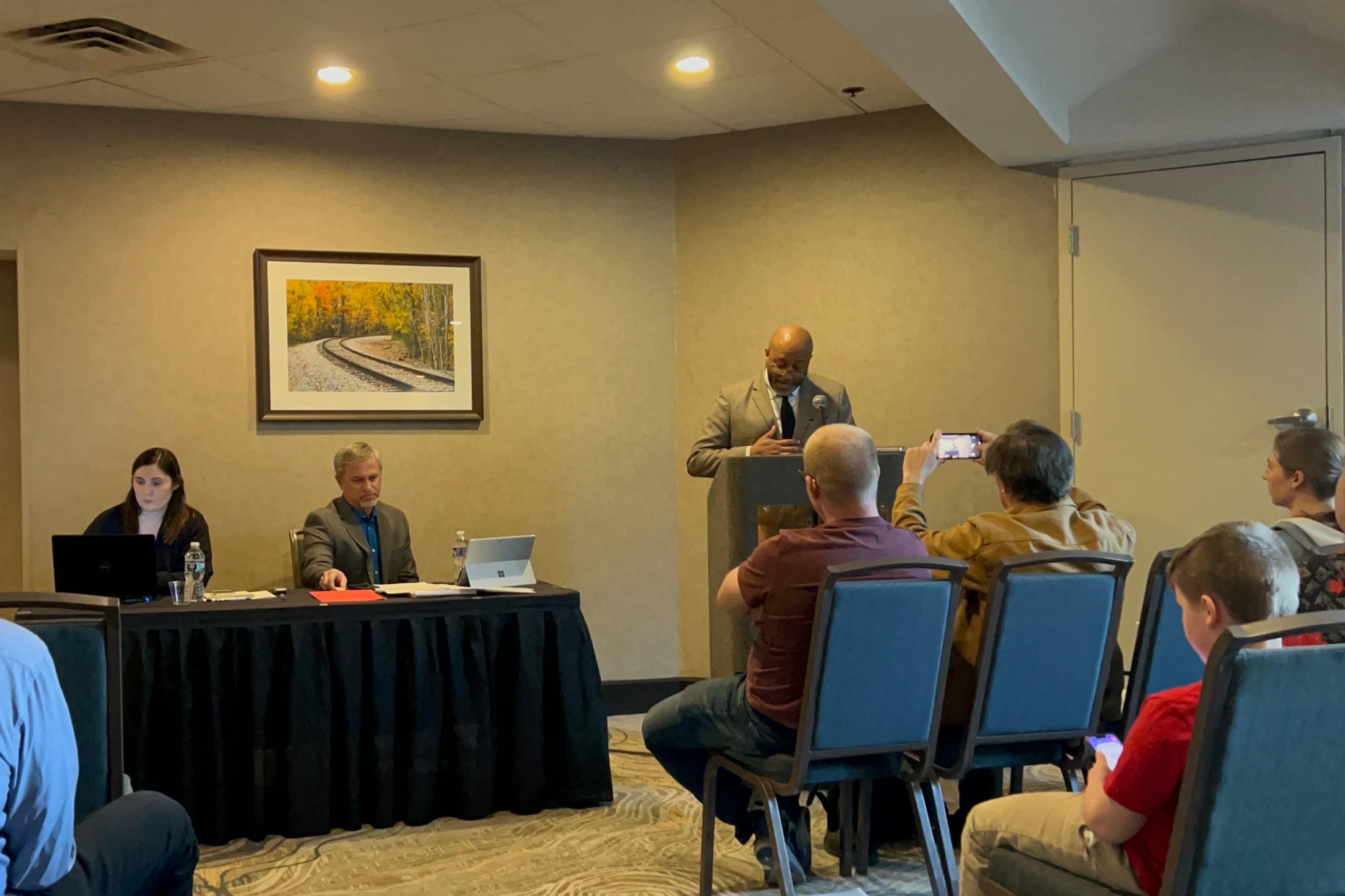 A man in a suit stands at a podium speaking before a crowd. Beside him are two people sitting behind a table also facing the crowd.