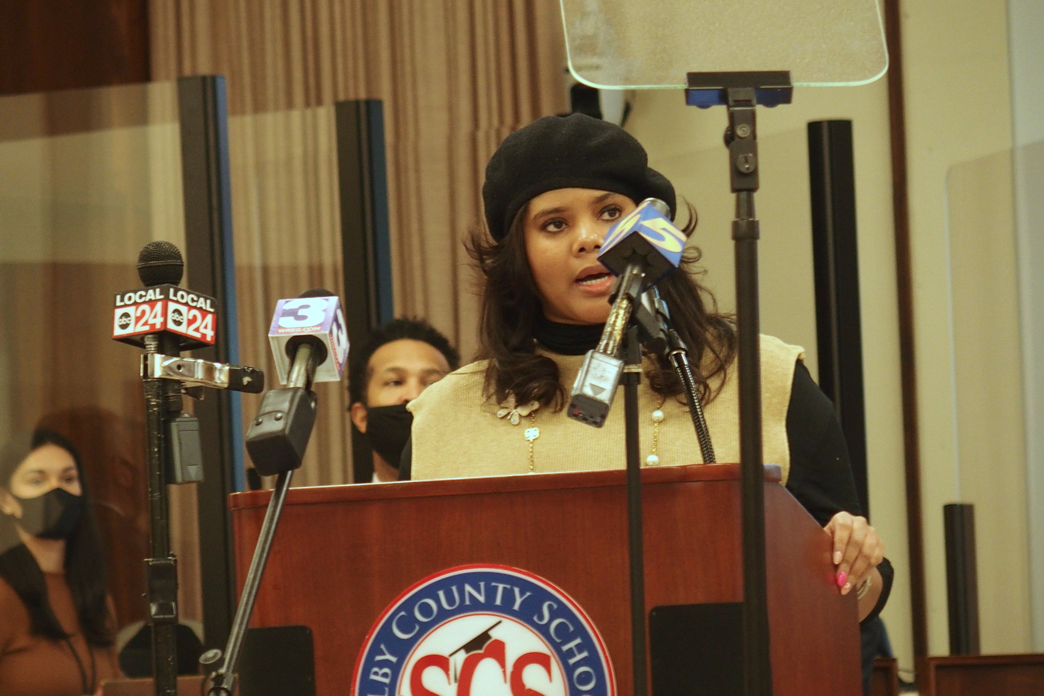 Miska Clay Bibbs stands behind a podium with TV stations microphones