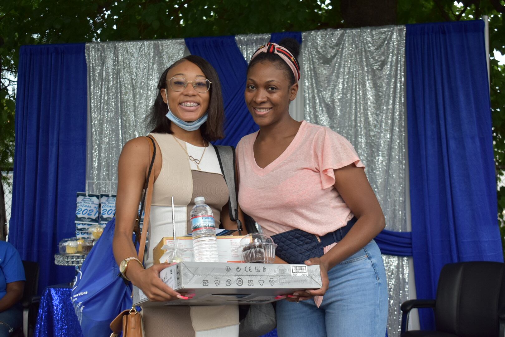 A girl wearing a tan dress holds a laptop box and a bag. She stands next to a woman wearing a pink shirt.