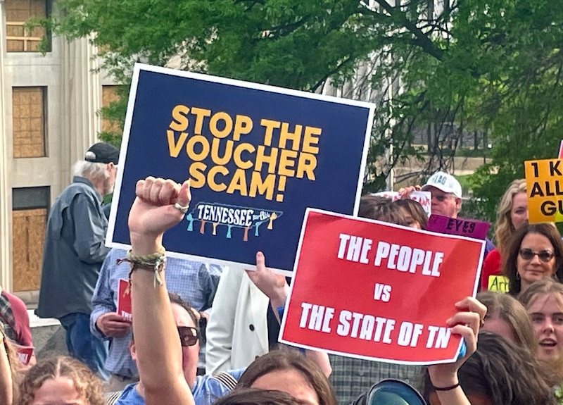 A close up of two signs that protestors are holding up outside with a group of people in the background.