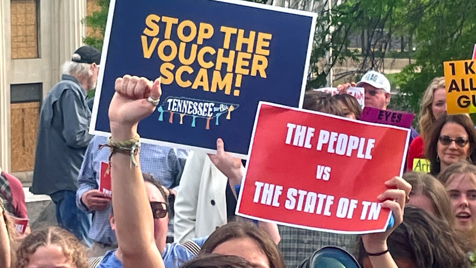 A close up of two signs that protestors are holding up outside with a group of people in the background.