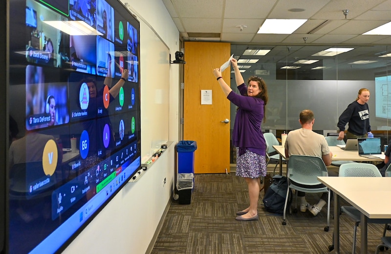 A woman in a purple jacket and dress holds up a piece of paper in a classroom.