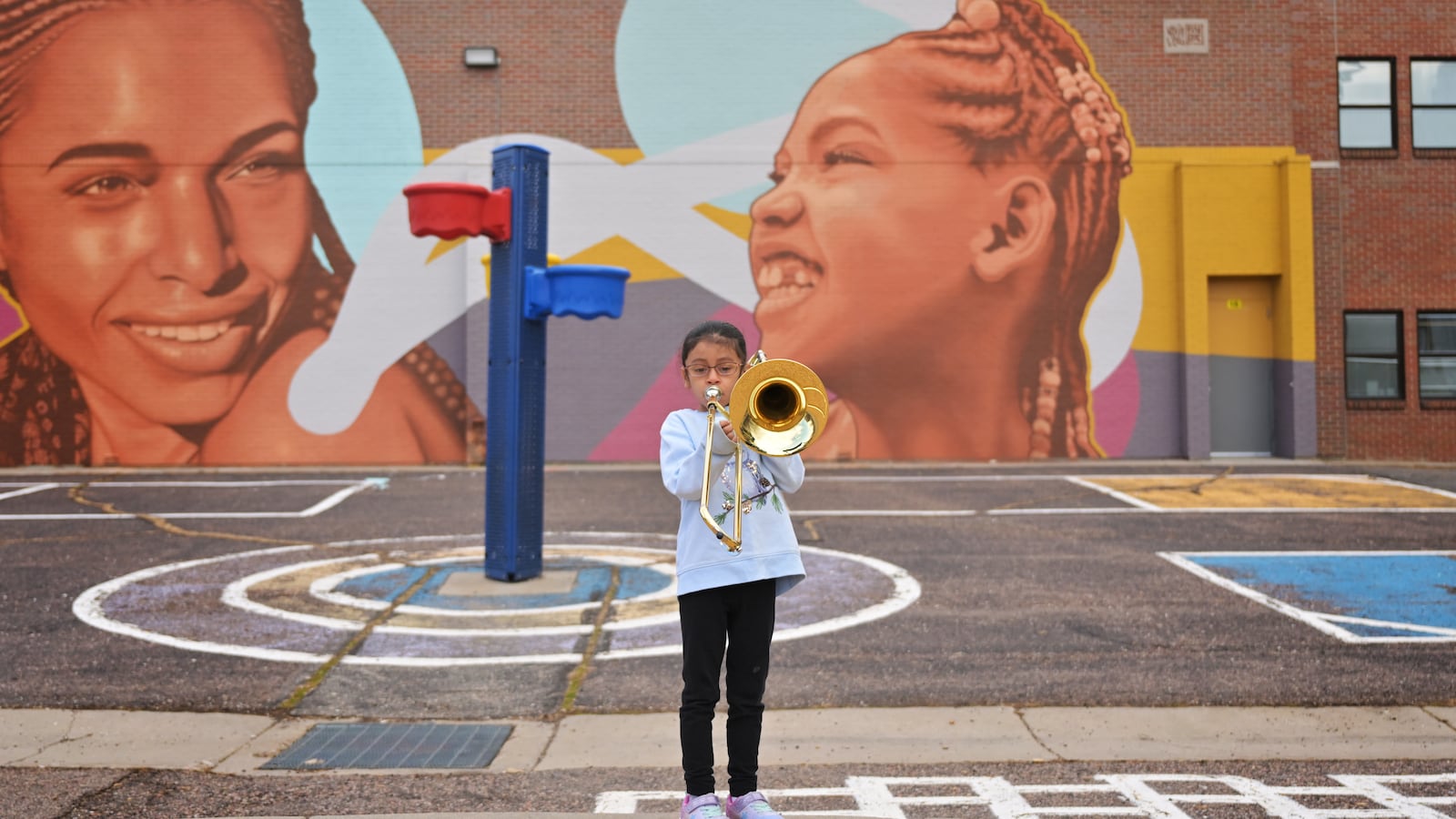 A young student plays the trombone on a sidewalk outside in front of a mural and playground.