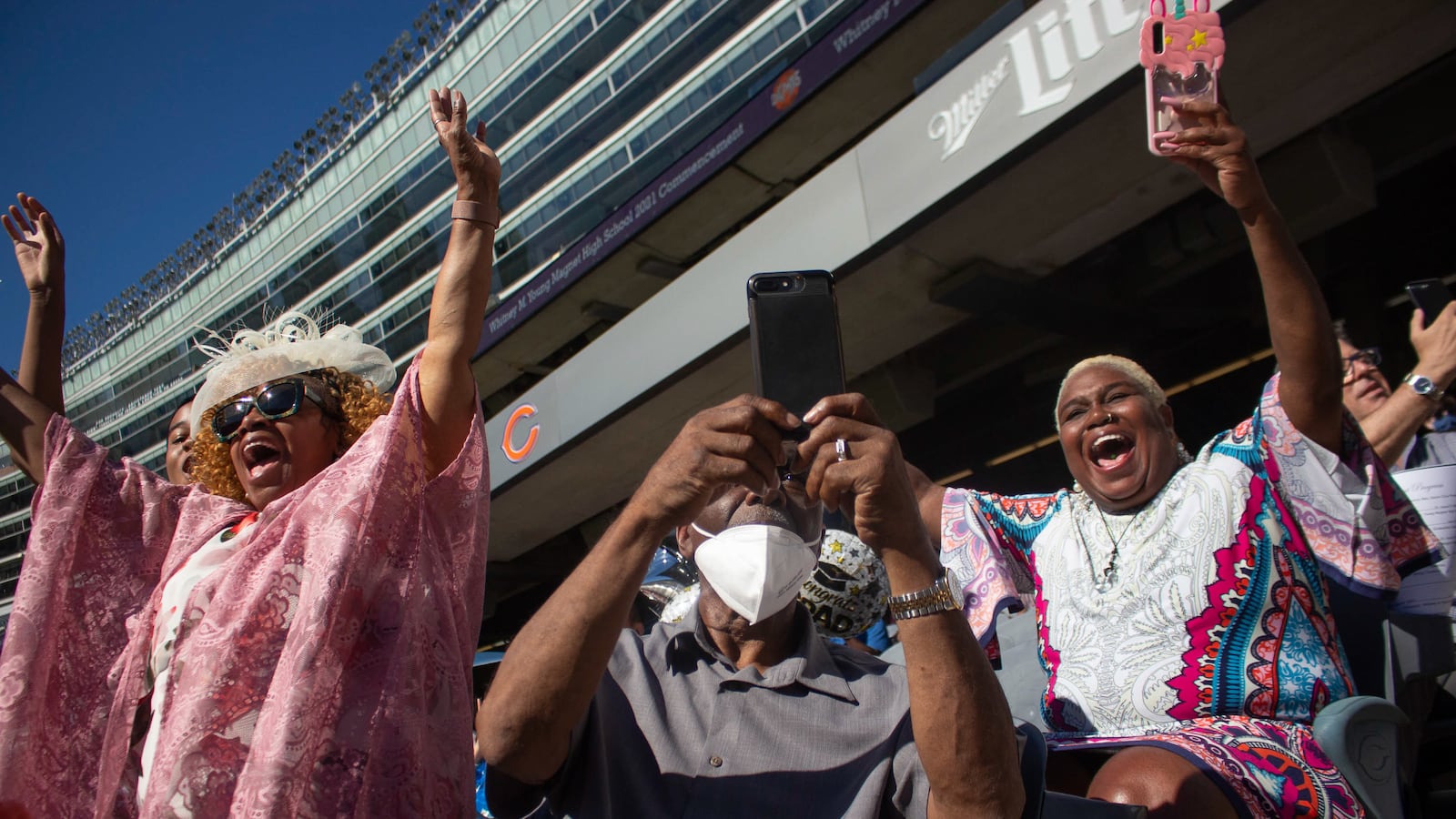 A woman wearing a pink shawl and sunglasses emphatically raises her arms as a man records on his cell phone, and a woman to the right wearing an intricate patterned dress excitedly cheers during a graduation ceremony.