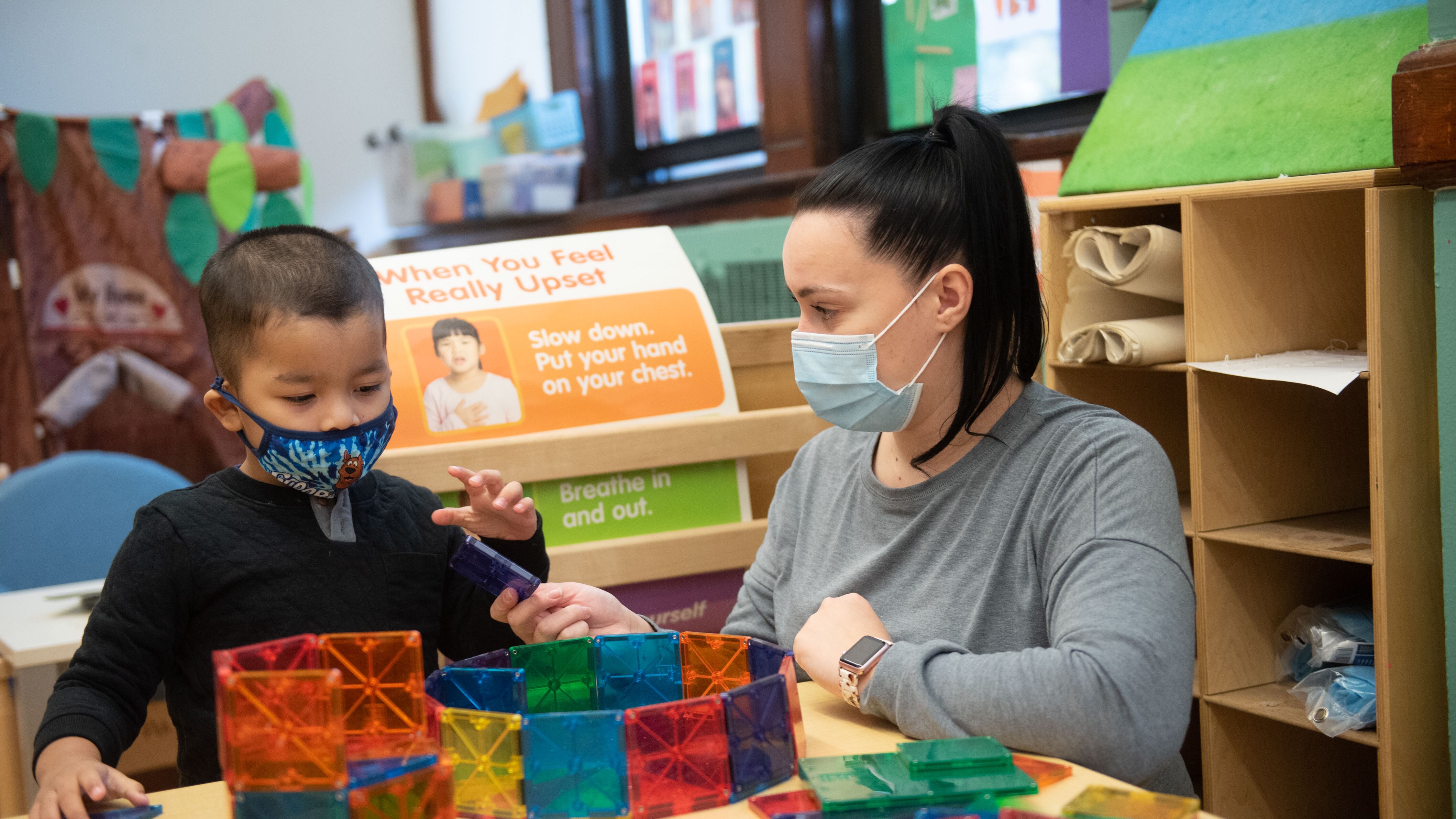 A preschool staff member works with a young boy with blocks in the classroom. They are both wearing protective masks.