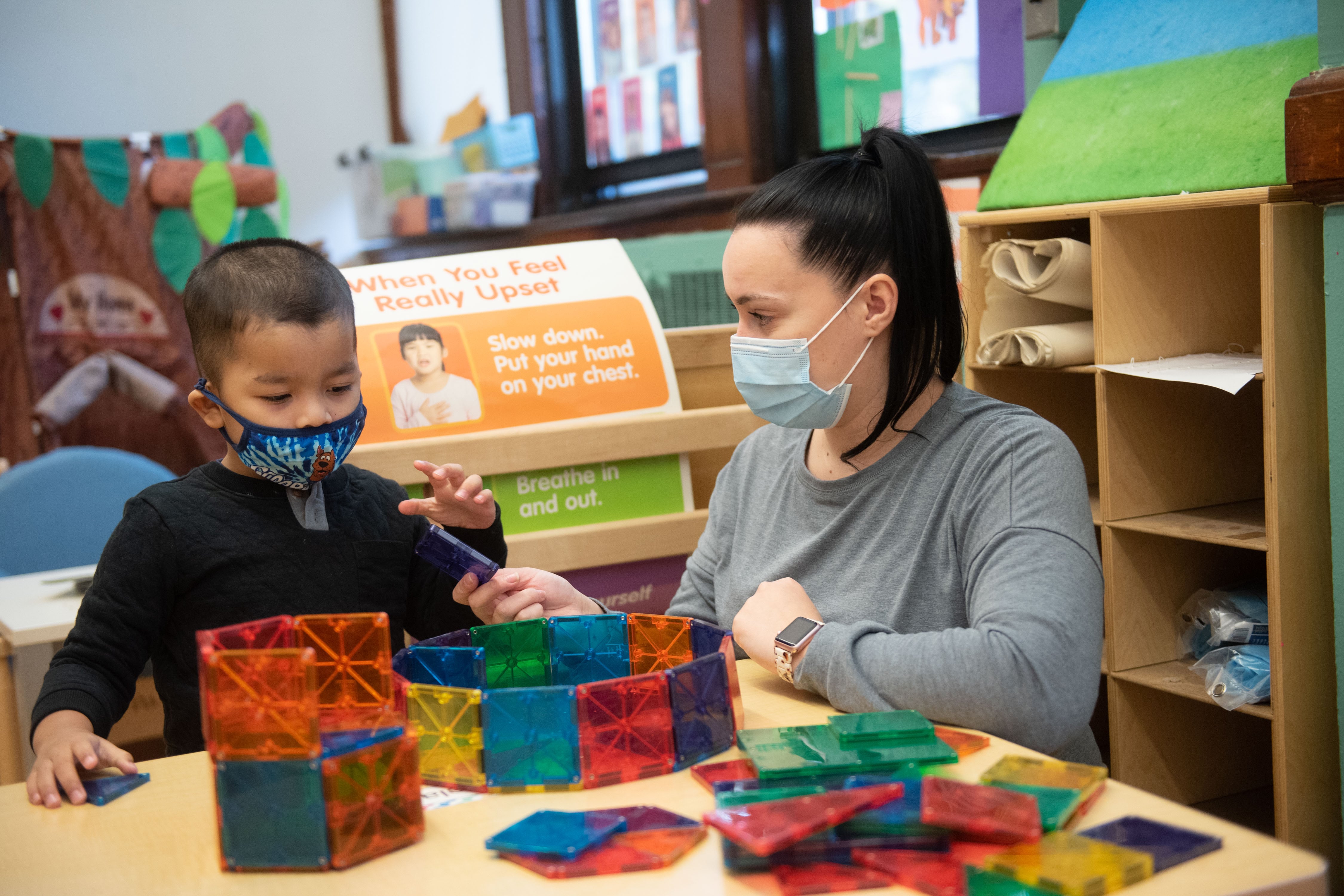 A preschool staff member works with a young boy with blocks in the classroom. They are both wearing protective masks.