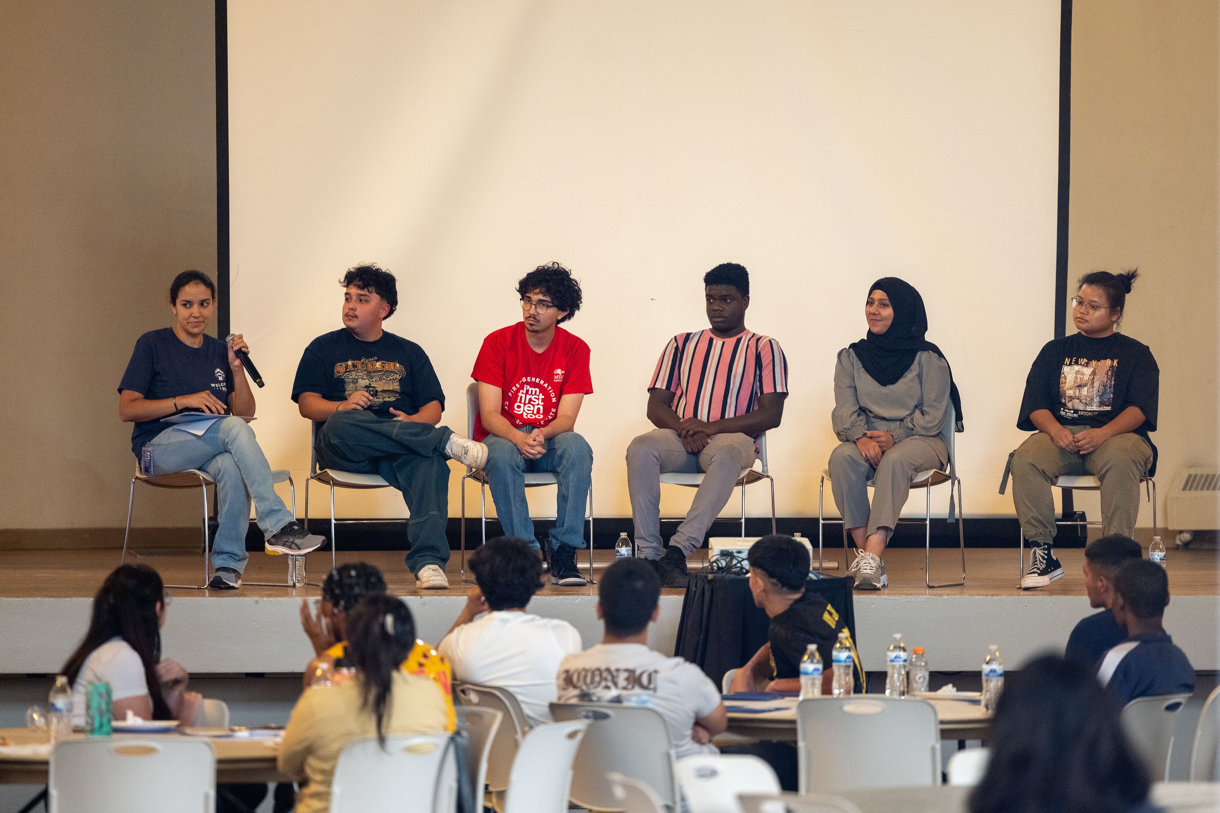 Six students sit in a line of chairs on a stage with people sitting in the foreground facing them in an auditorium.