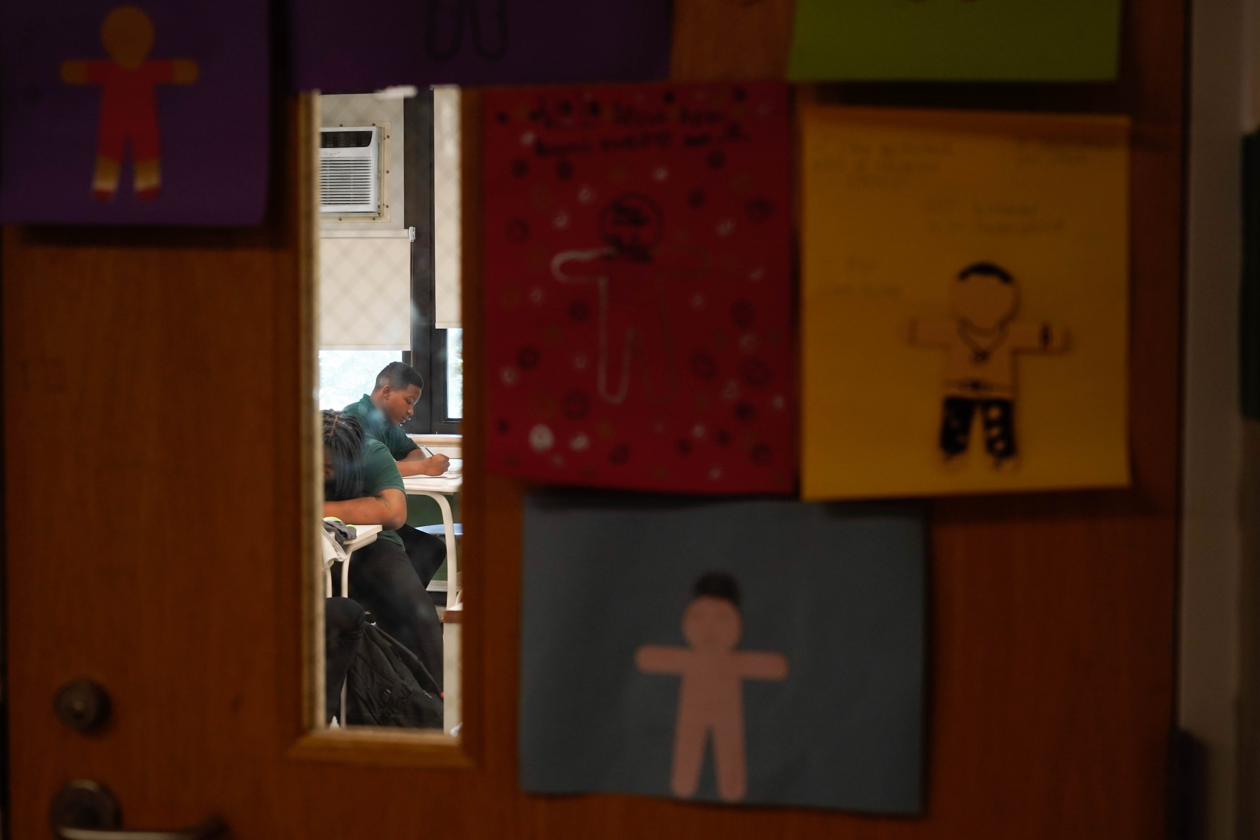 Seen through a small glass window into a classroom a young boy sits at a desk.
