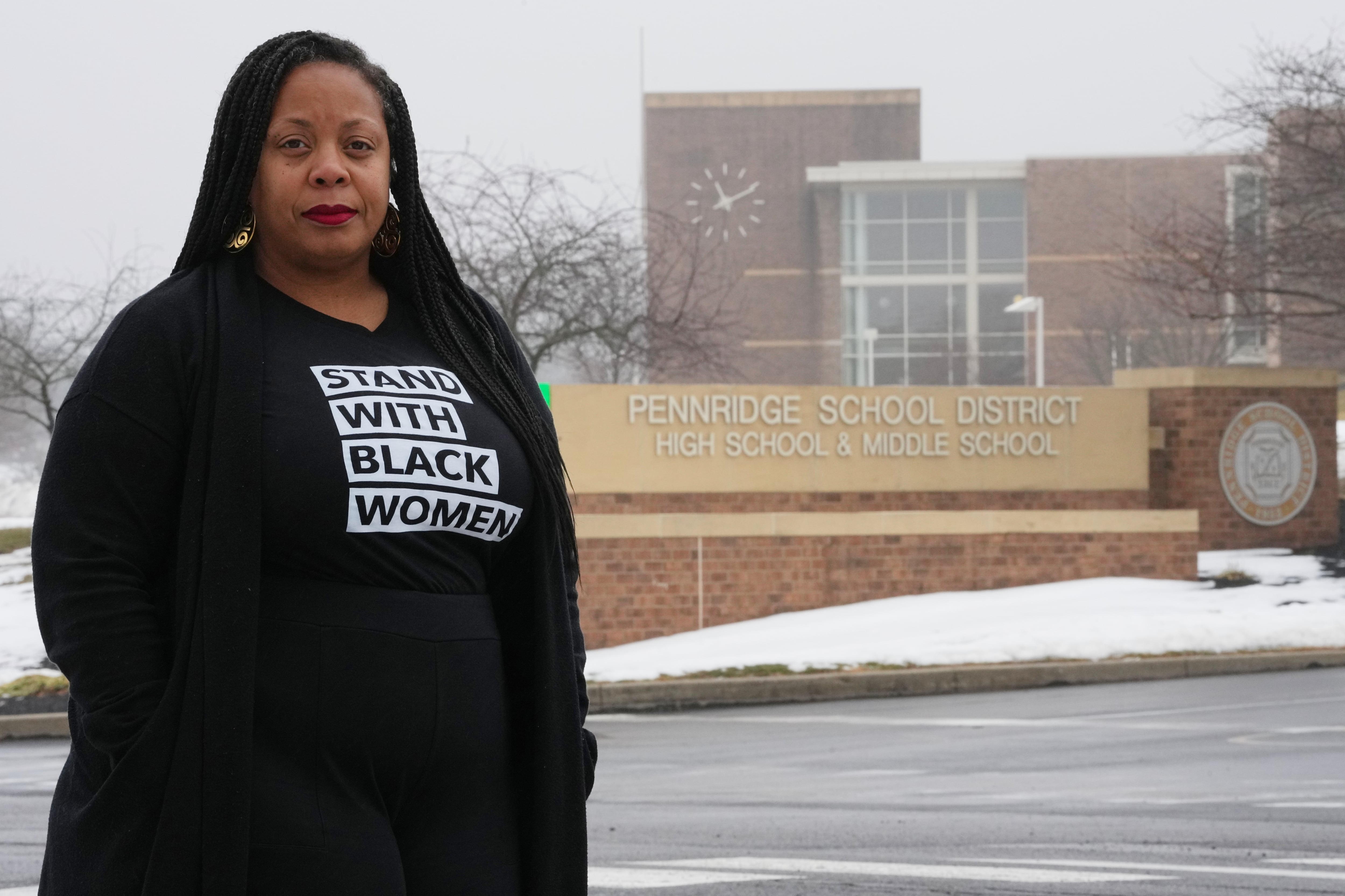 A Black woman stands outside a school building in light snow in the winter. Her face is serious. She has long hair and is dressed in black. Her t-shirt reads "Stand with Black Women."