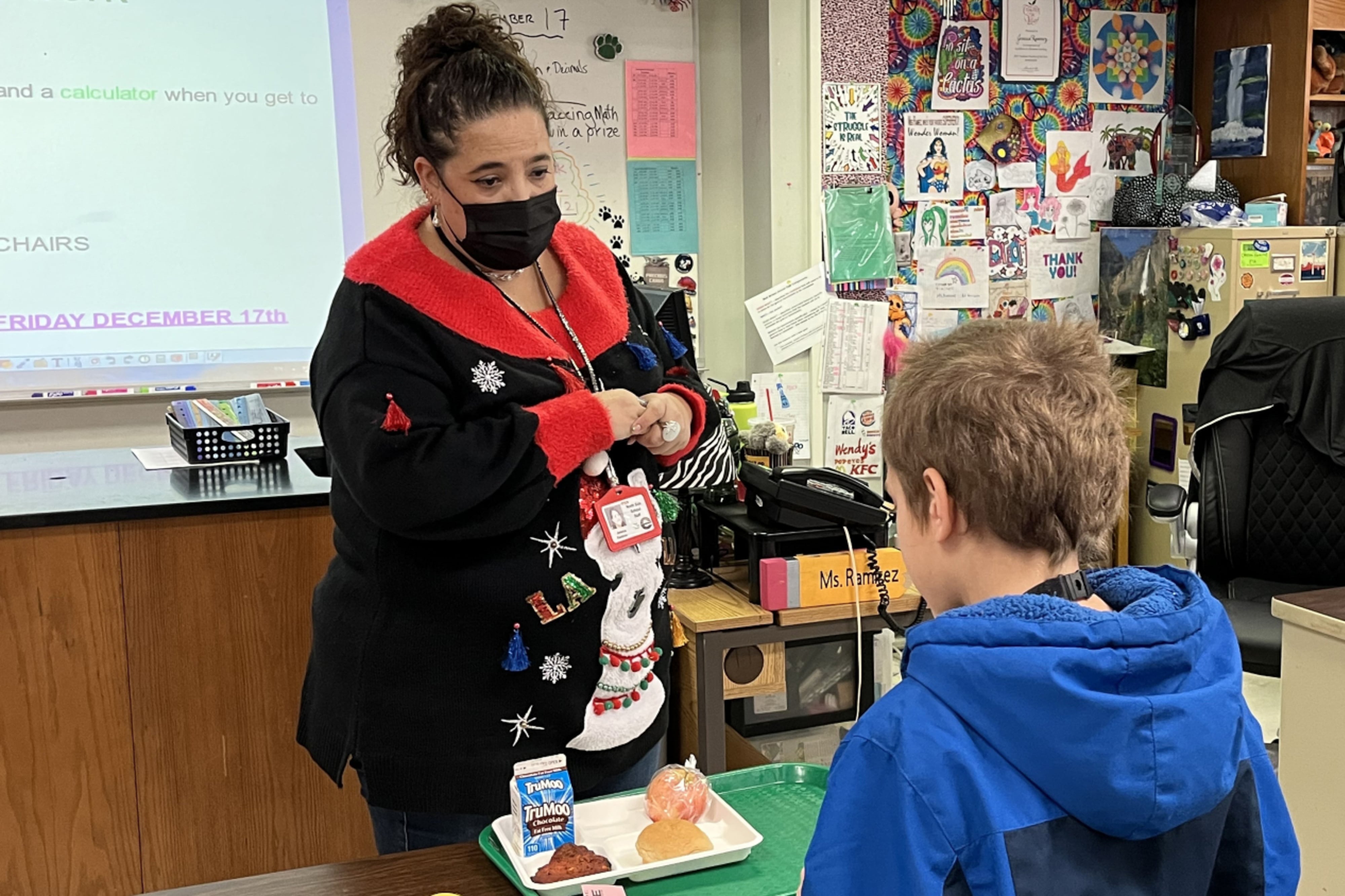 A woman wearing a mask and in a black and red shirt stands in front of a desk while speaking to a child in a blue hoodie sweatshirt.