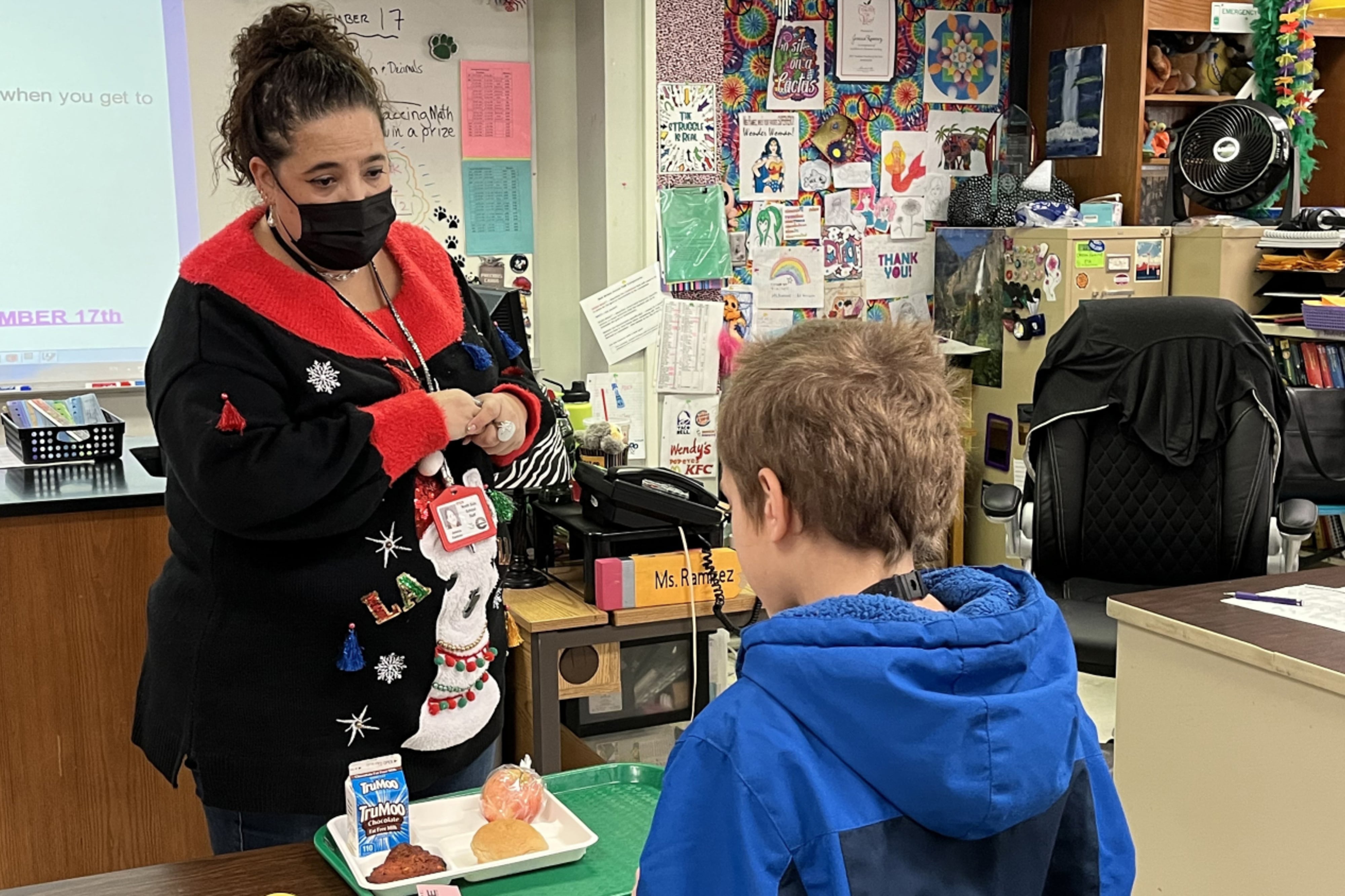 A woman wearing a mask and in a black and red shirt stands in front of a desk while speaking to a child in a blue hoodie sweatshirt. 