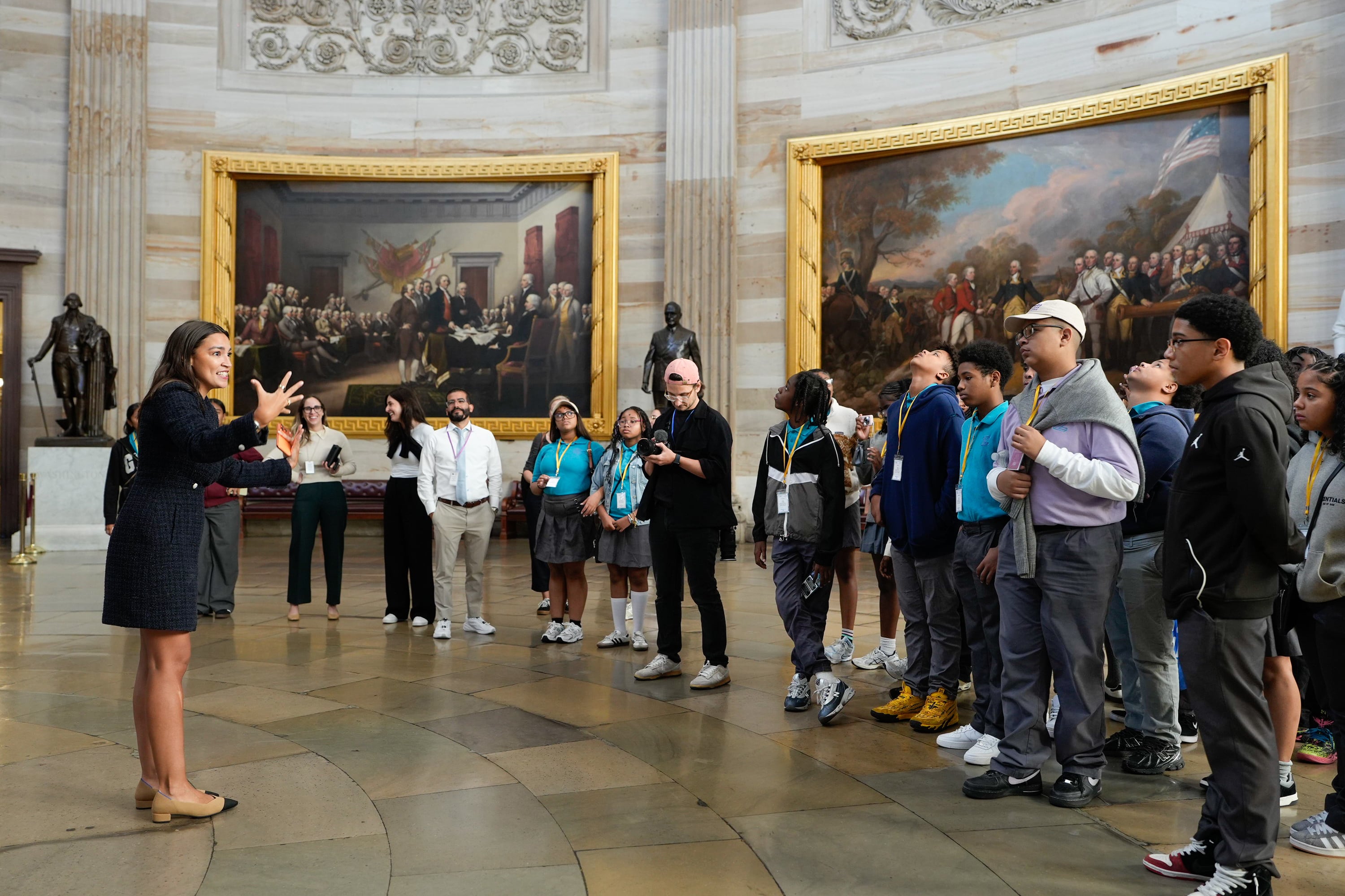 A woman in a dark suit stands at left in front of a group of students and two giant paintings.