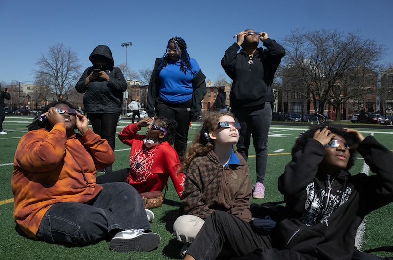 A group of high school students all wearing eclipse viewing glasses look up at the sky outside on a sports field.