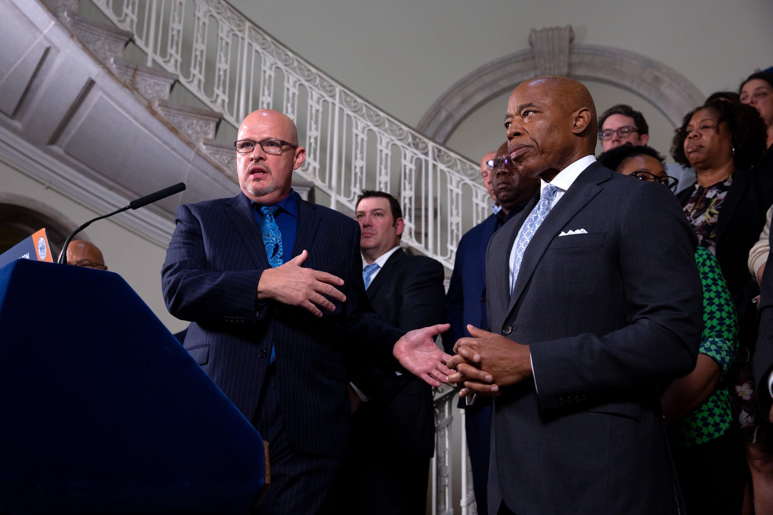A man in a suit with a dark blue tie stands at a podium next to a man in a suit with a pale blue tie.