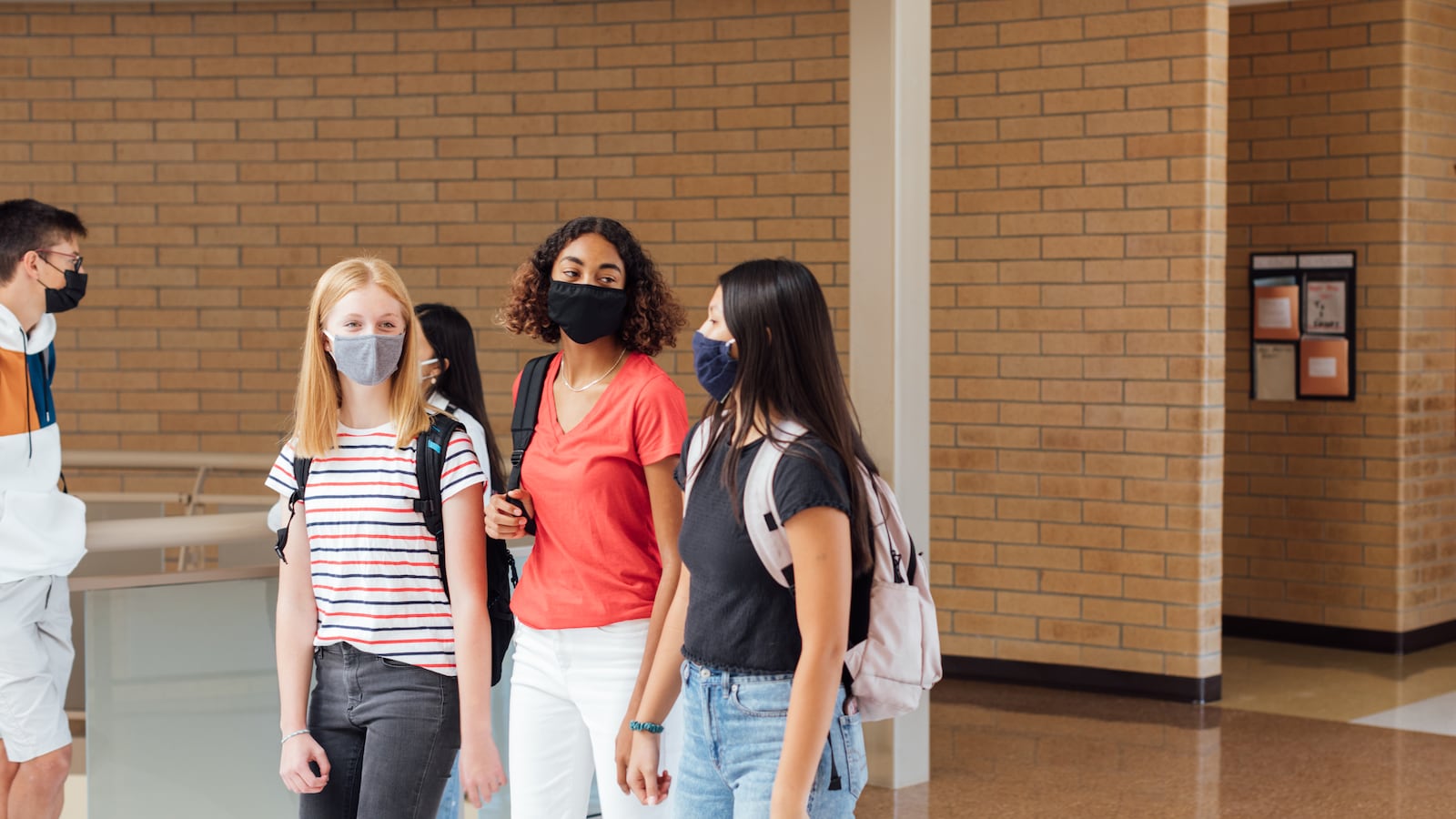 Three teenage girls walk through the hallway at their high school. They are wearing facing masks.