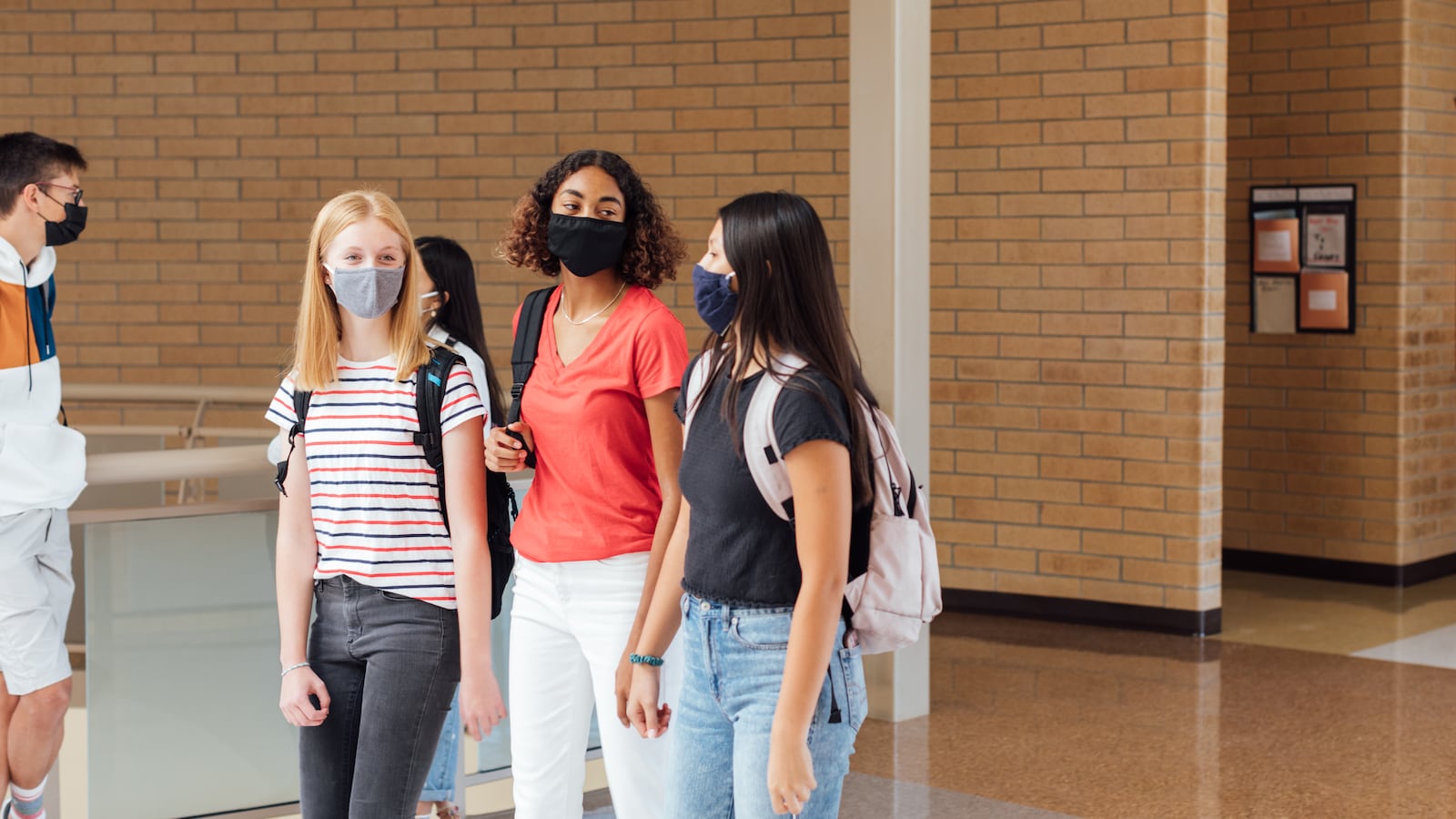 Three teenage girls walk through the hallway at their high school. They are wearing facing masks.
