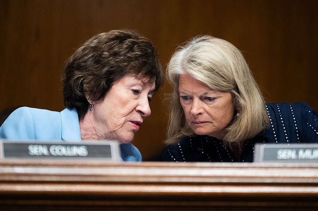 Senators Susan Collins, a white woman with short dark hair and wearing a light blue blazer, and Lisa Murkowski, a white woman with shoulder-length sandy-colored hair wearing in a navy pinstripe blazer, lean their heads together to talk while seated at a dais. Their name plates are visible in the foreground.