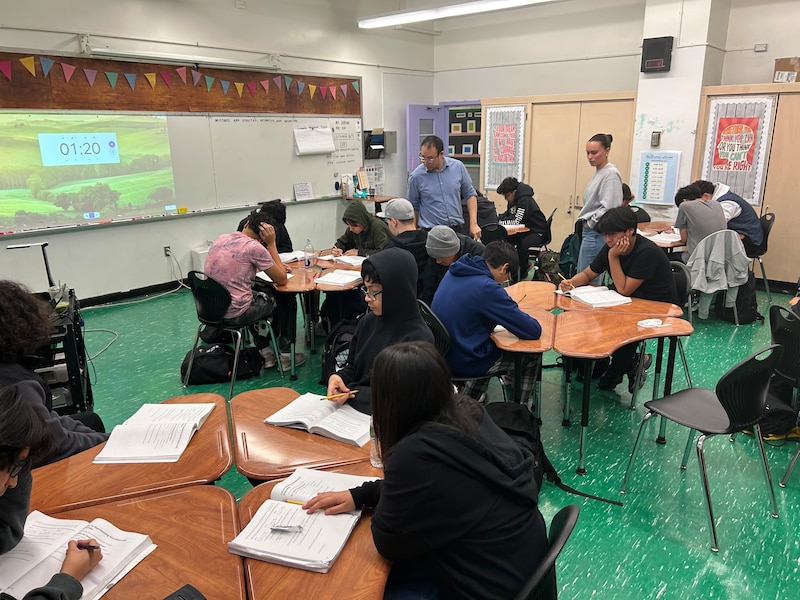 A classroom with students sitting at desks pushed into a circle. An adult man and an adult woman are standing, checking on student work. The classroom has a green floor.