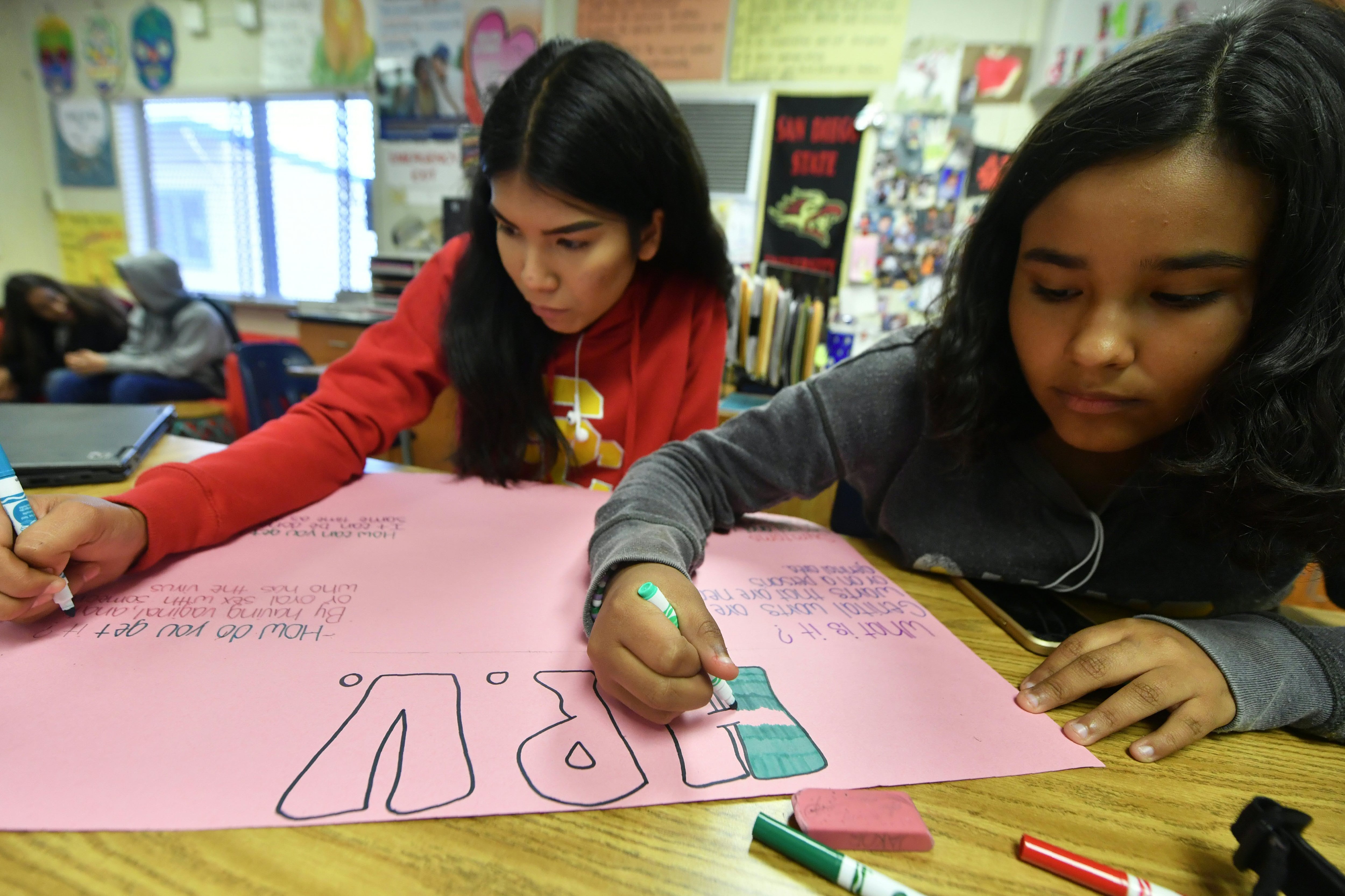 A girl in red and a girl in gray create a poster on pink board.
